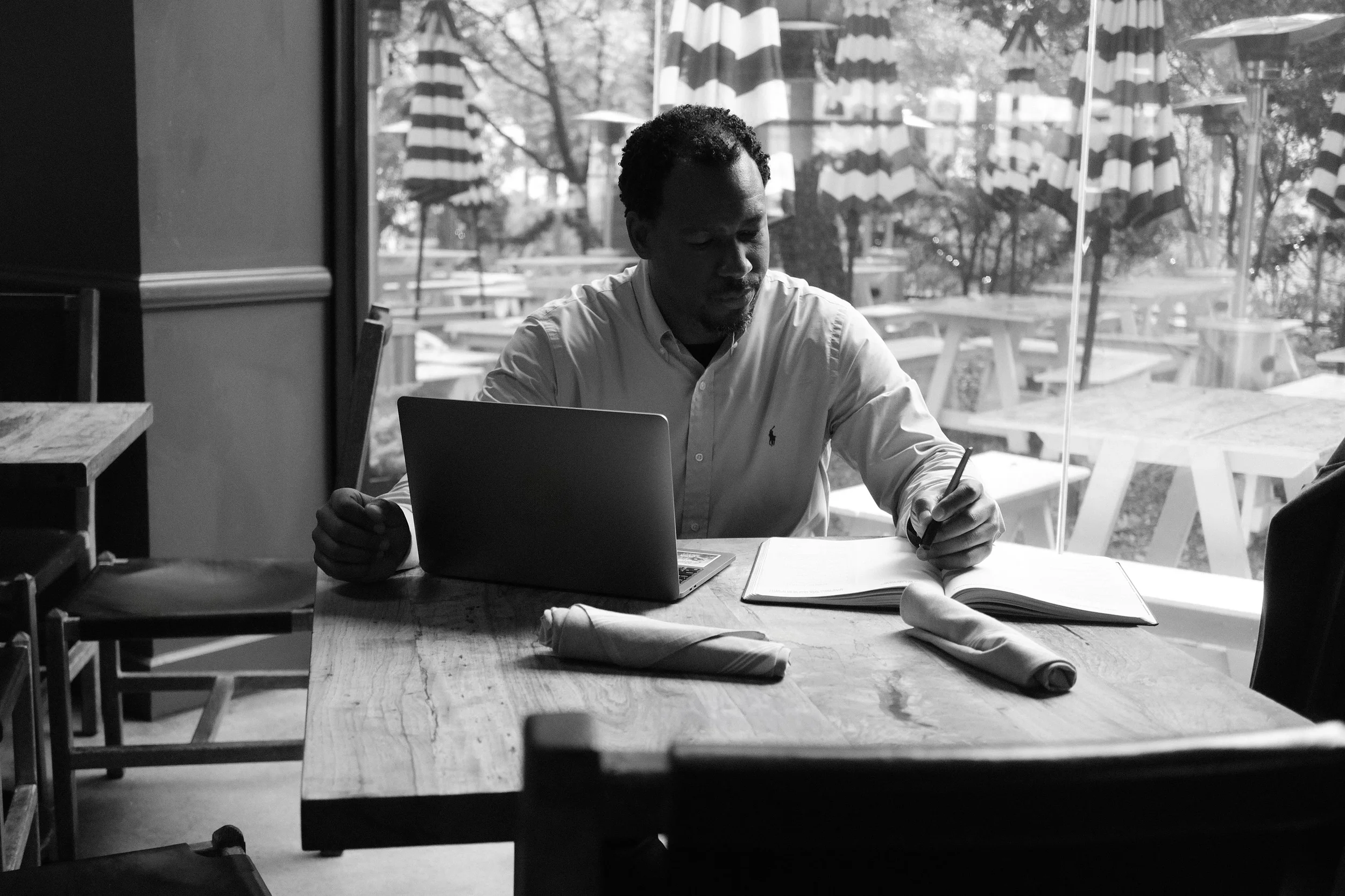 Black and white photo of a professional man working on a laptop and writing in a notebook at a wooden table inside a cafe.