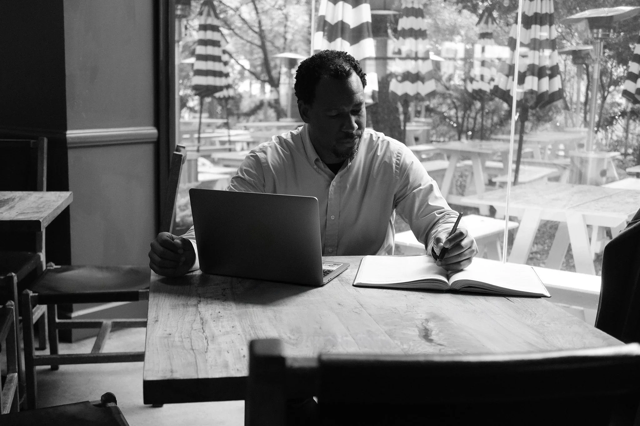 Black and white photo of a professional man working on a laptop and writing in a notebook at a wooden table inside a cafe.