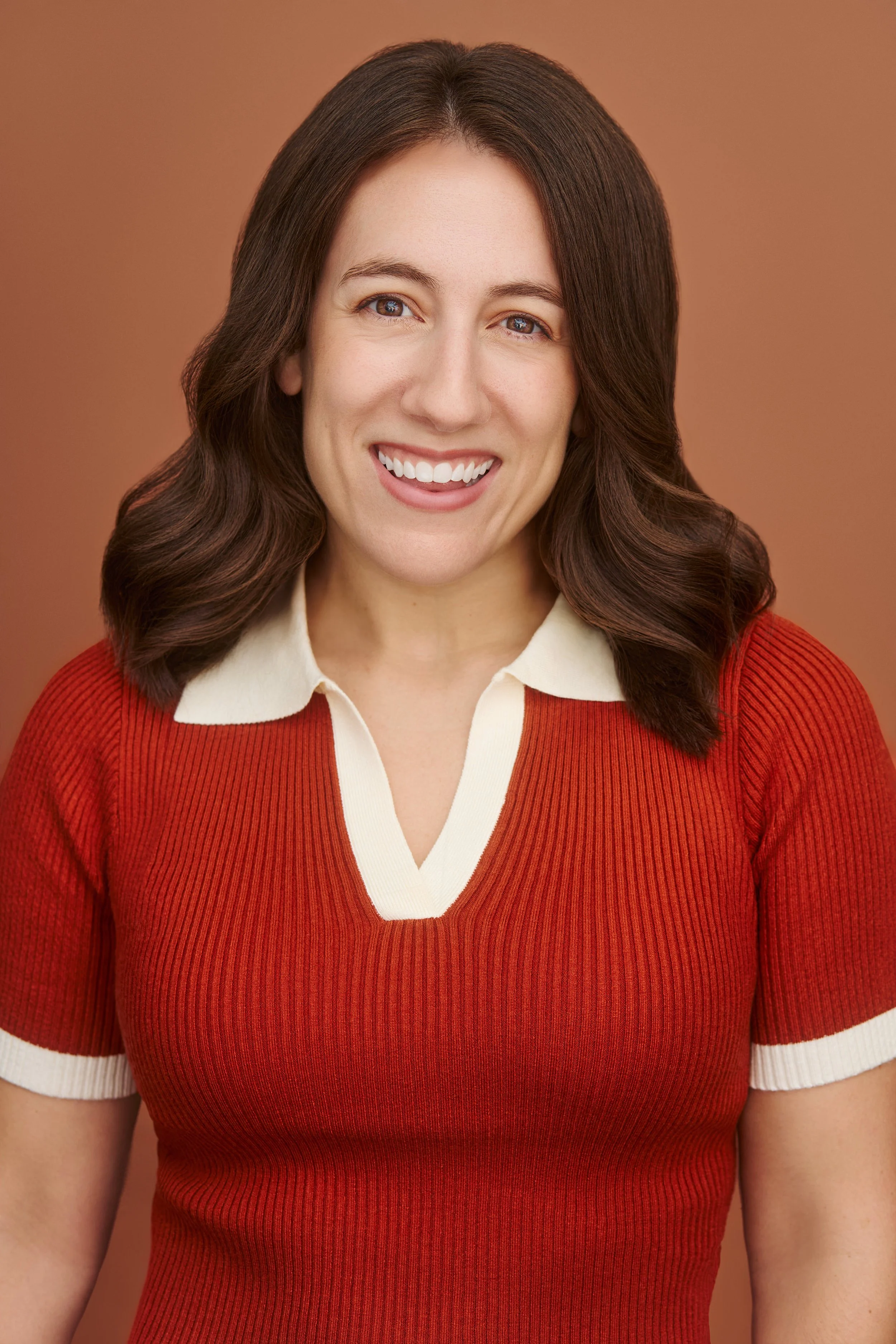 A woman with medium-length wavy dark brown hair smiling, wearing a red and cream-colored collared sweater against a solid light brown background.