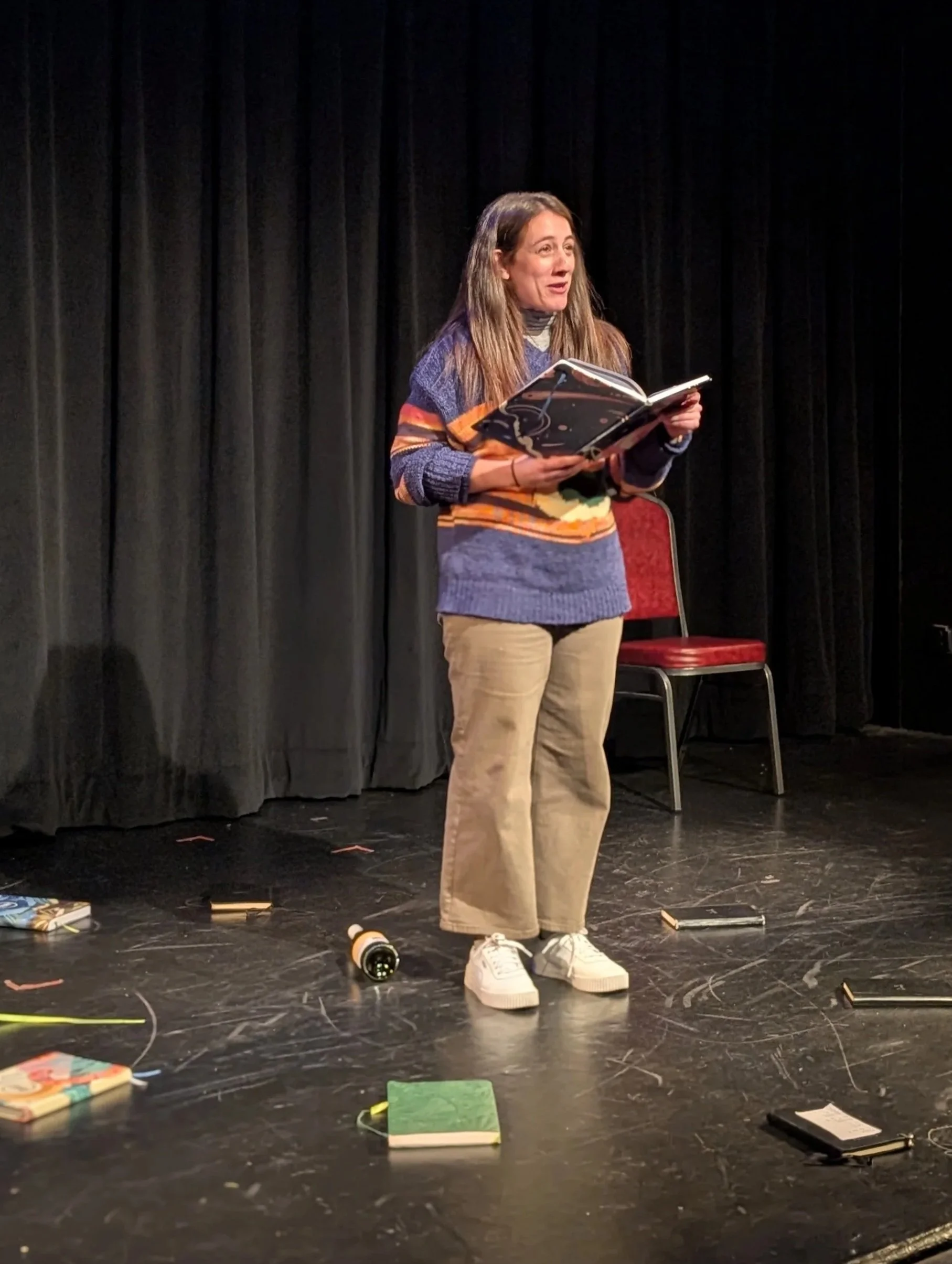A woman standing on a black stage with a black curtain backdrop, holding a large book or script, wearing a colorful sweater, beige pants, and white sneakers, surrounded by scattered notebooks and objects, with a red chair behind her.