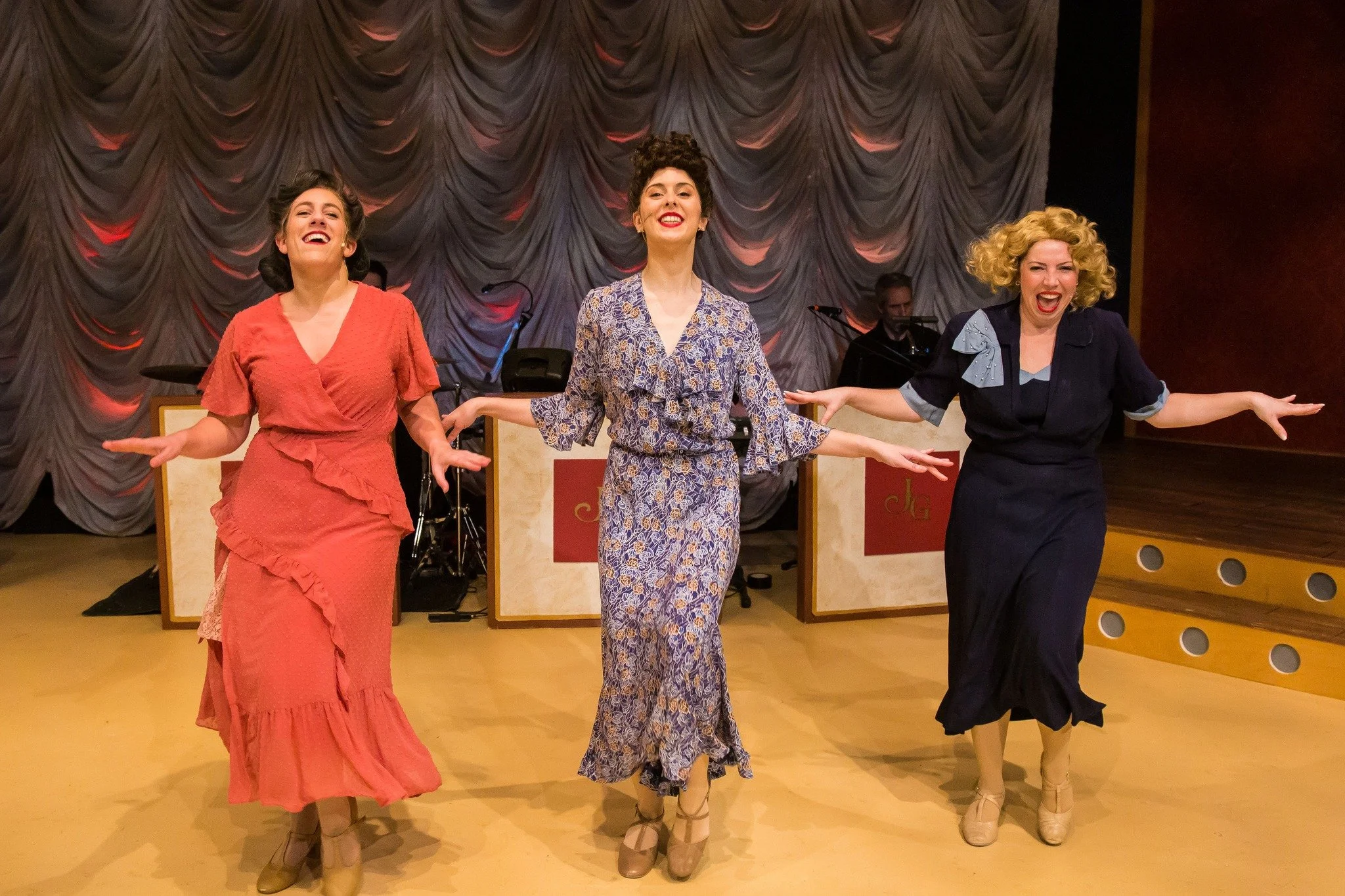 Three women in vintage dresses dancing with arms outstretched on stage, with a band in the background.