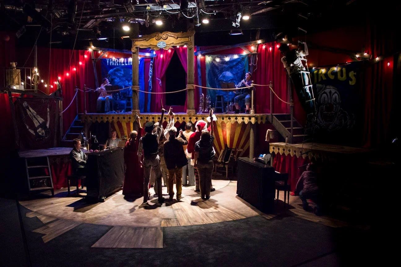 A small circus stage with red curtains and golden decorations, featuring two performers playing string instruments, and an audience of people gathering in front, some wearing Santa hats.