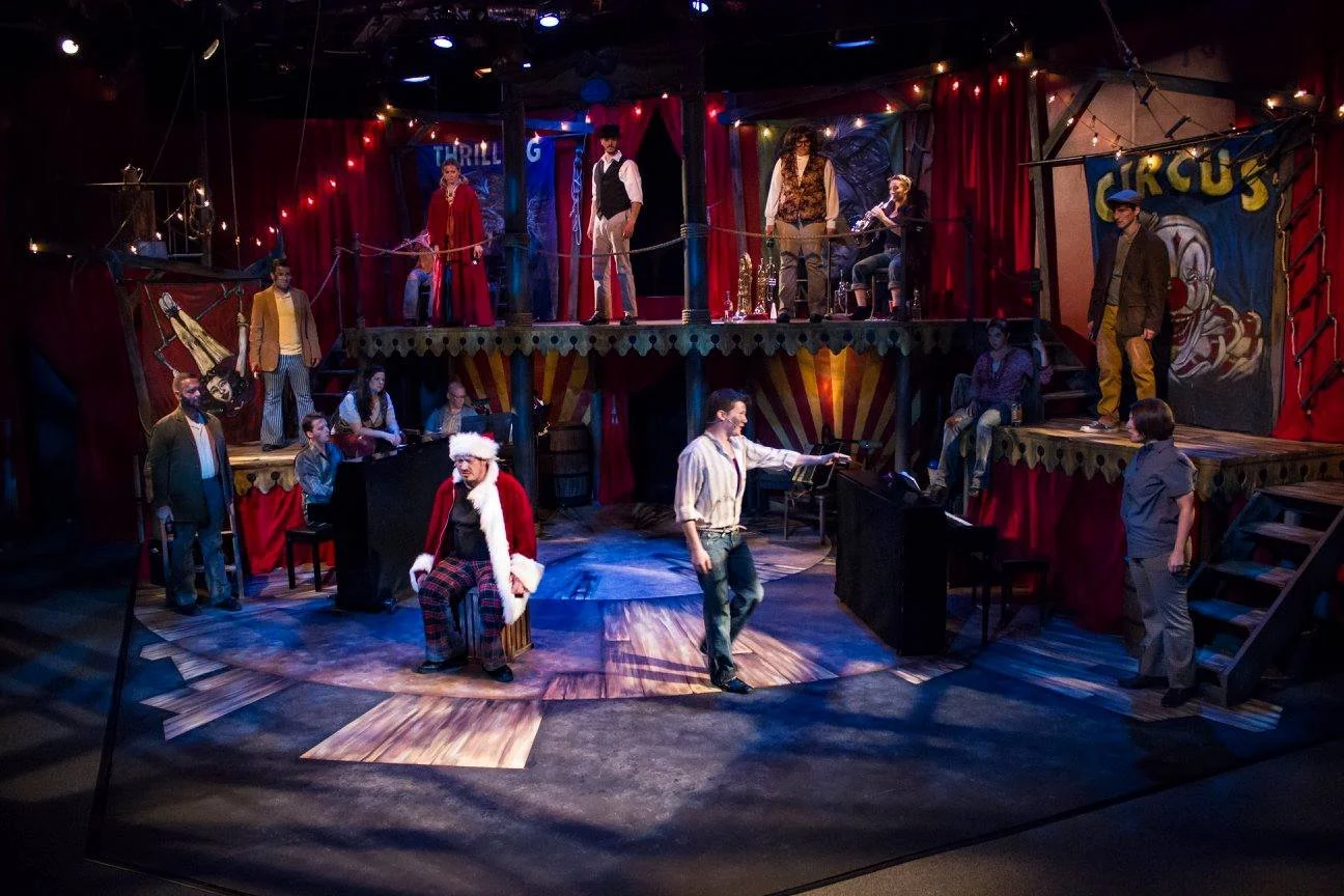 Actors on a circus stage with red curtains, festive lights, and circus banners, rehearsing or performing.