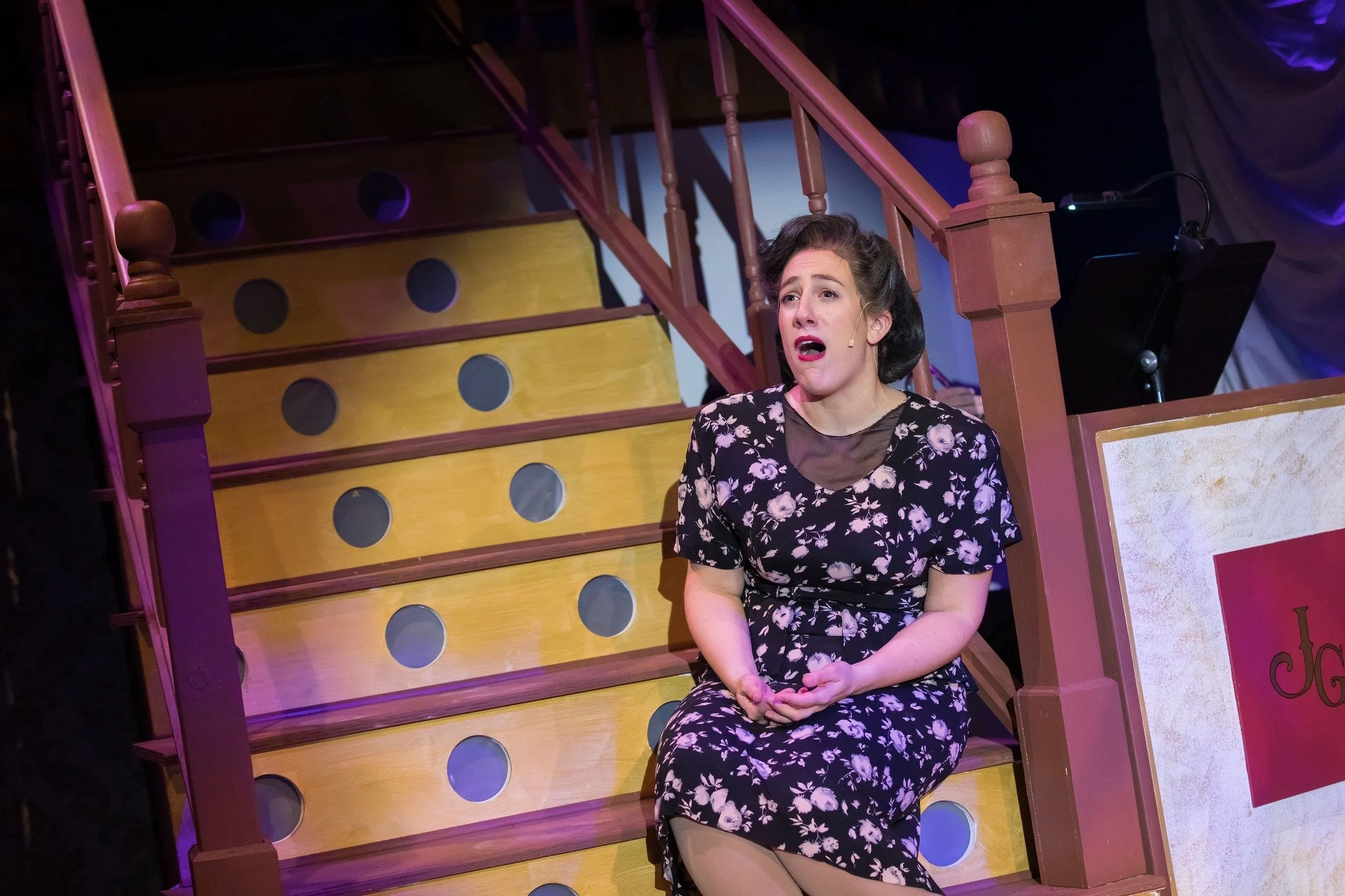 A woman with dark hair sitting on a staircase, singing with an emotional expression, wearing a black floral dress.