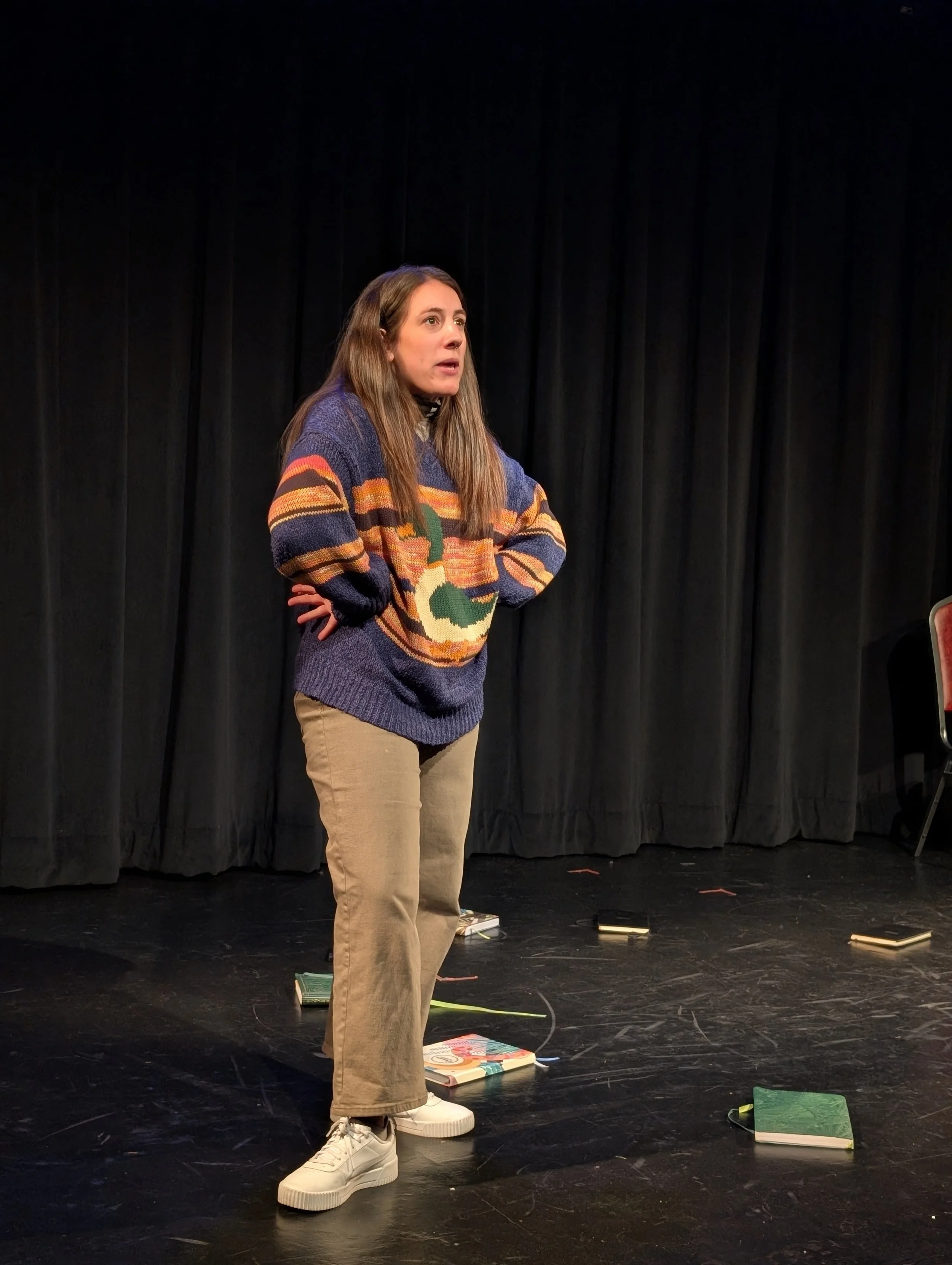 A woman with brown hair stands on a stage with black curtains, wearing a colorful sweater, beige pants, and white sneakers. There are scattered books on the floor.