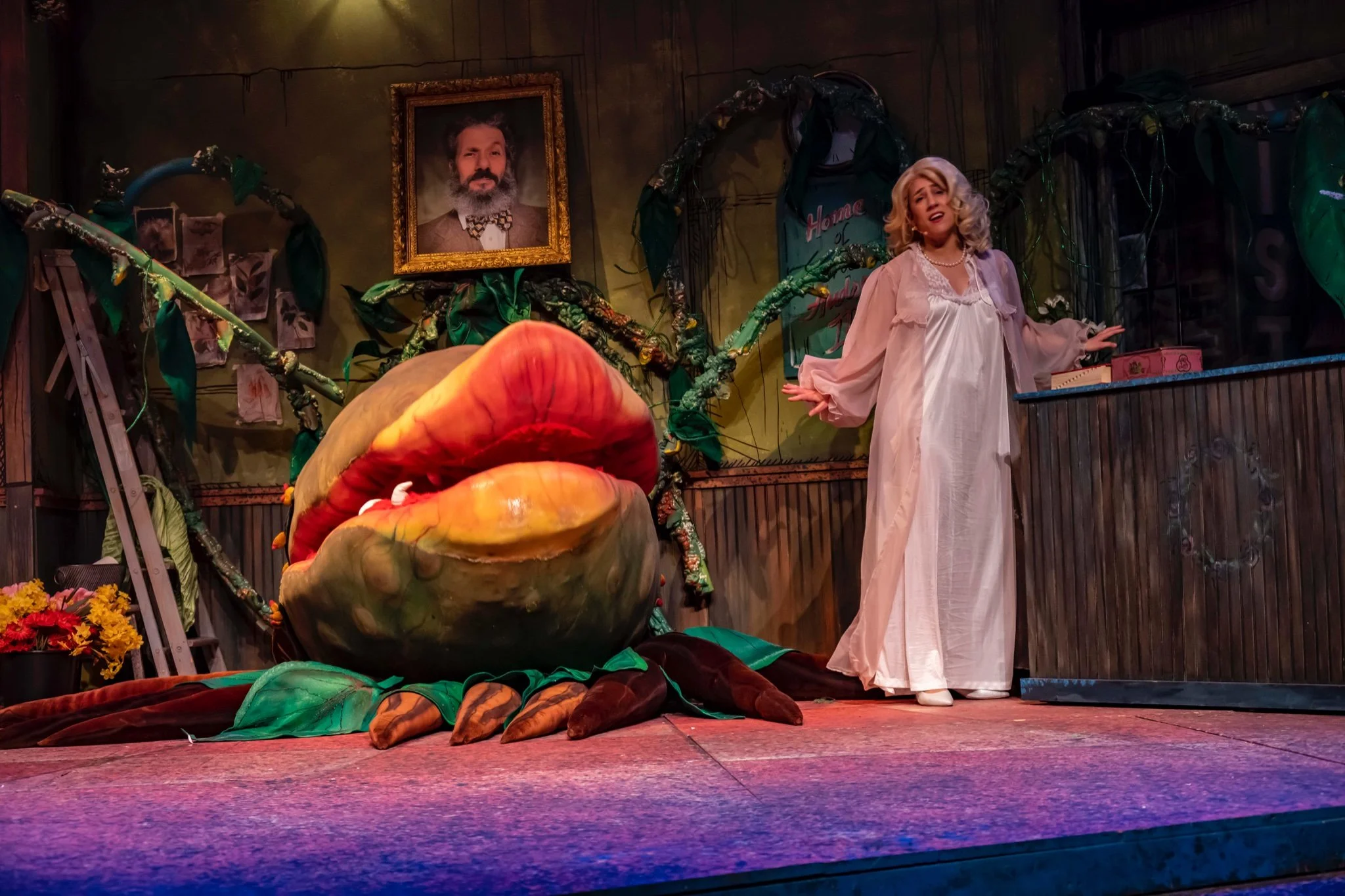 A theatrical scene with a woman in a long white gown standing behind a wooden counter, a large puppet of a mouth with big lips and teeth on the stage, and a framed portrait of a man hanging on the wall in the background. The stage is decorated with g