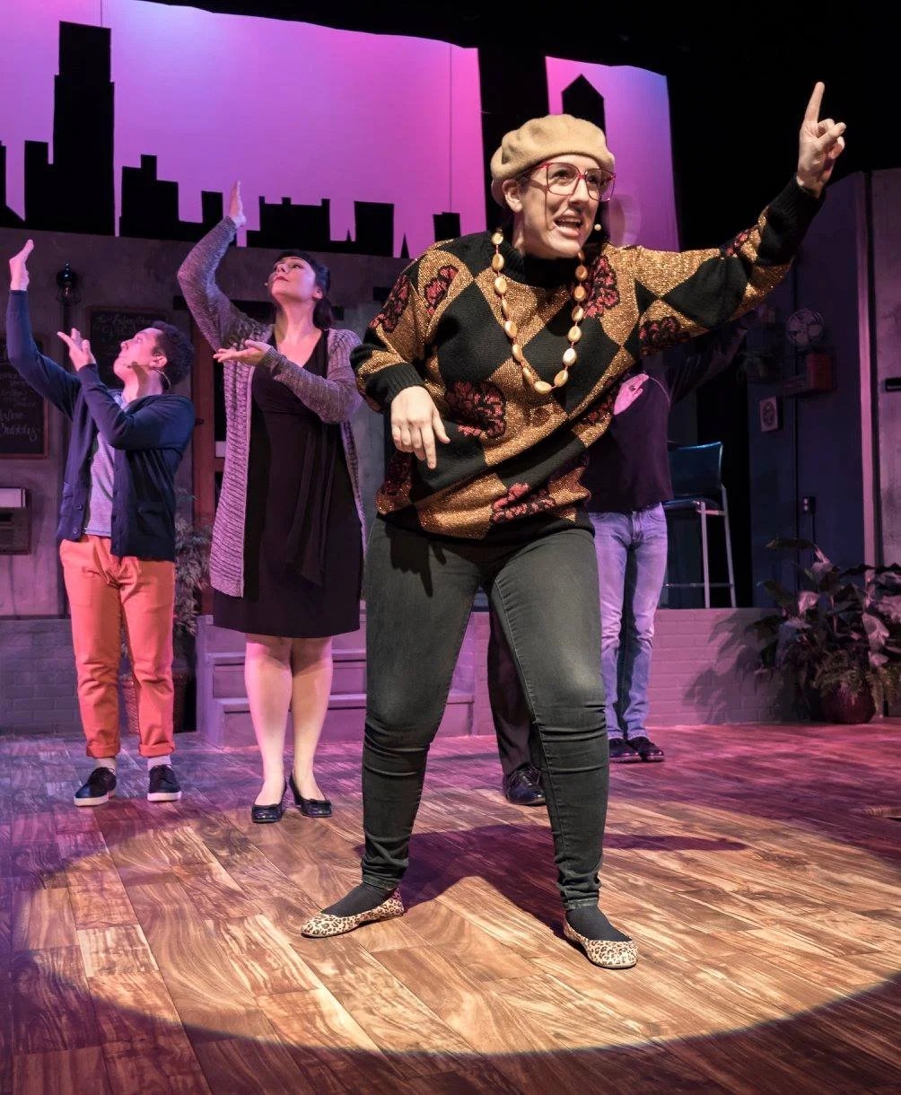 The image shows a stage performance with five actors. The woman in the front is speaking or singing passionately, wearing a beige beret, glasses, a colorful patterned sweater, and leopard print shoes. The other actors in the background are also expre