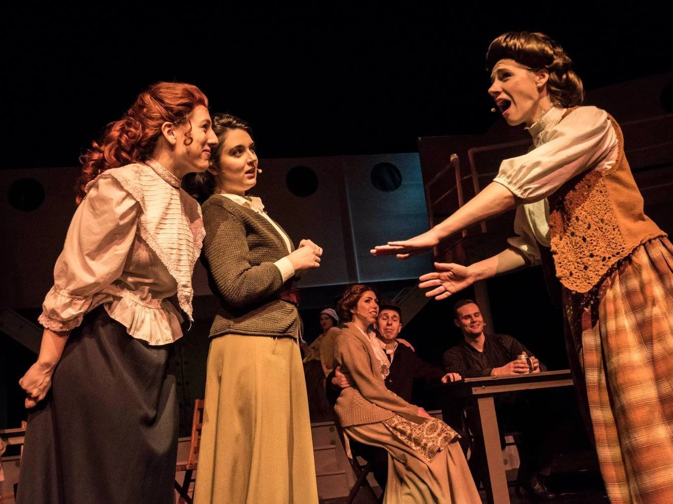 Four women on stage engaged in a theatrical performance, with two men seated in the background.
