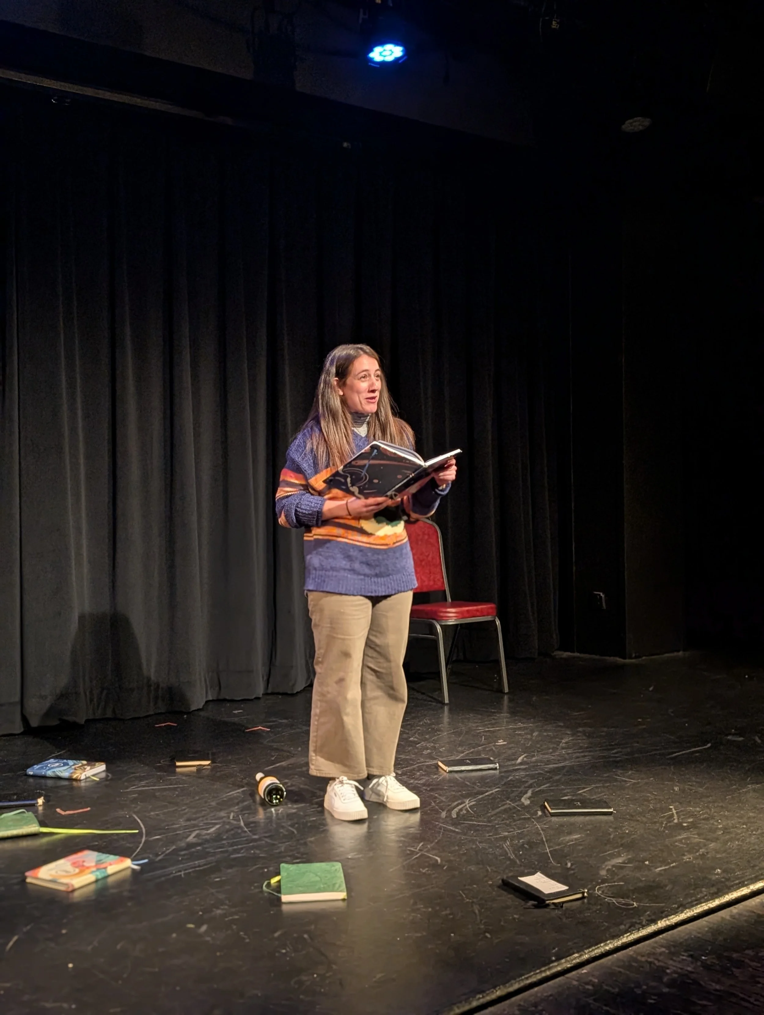 Woman on stage reading from a book with scattered books and notebooks around her, black curtains in the background, and a red chair behind her.