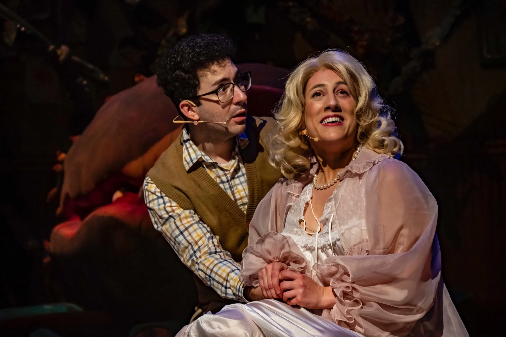 Two actors on stage performing a scene from a play. The man has curly dark hair, glasses, and a microphone, and is looking at the woman with a worried expression. The woman has blonde curly hair, is wearing a pearl necklace, and is looking upward wit