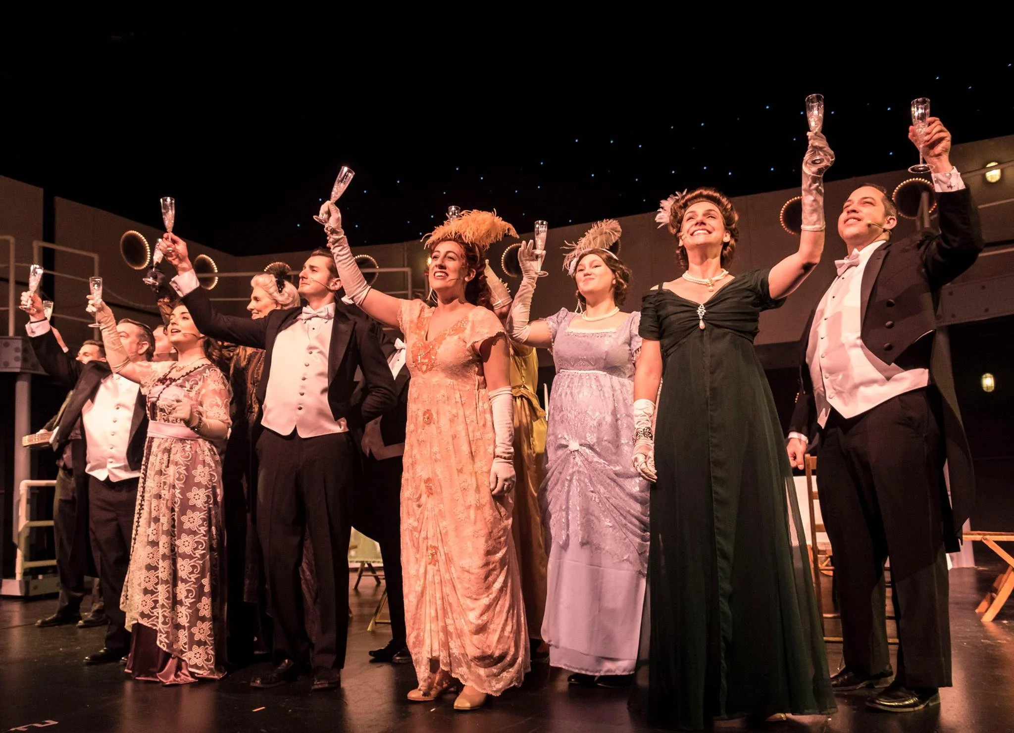 Actors on stage dressed in vintage 19th-century clothing, holding glasses of champagne in a party toast, celebrating after a theatrical performance.