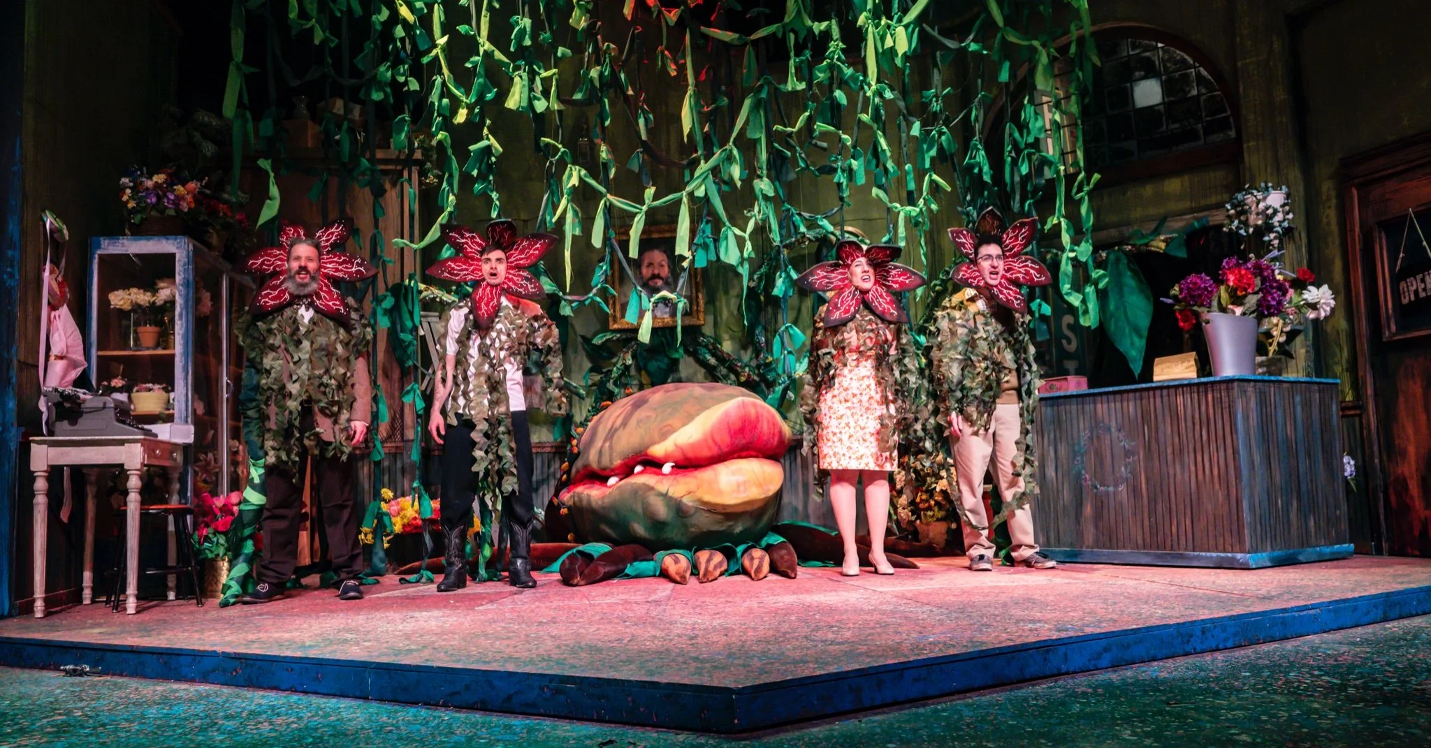 Stage scene with six performers dressed as flowers and plants, surrounded by greenery and flowers, with a large, whimsical plant or creature at center stage, and props like a desk, shelves, and flower pots.