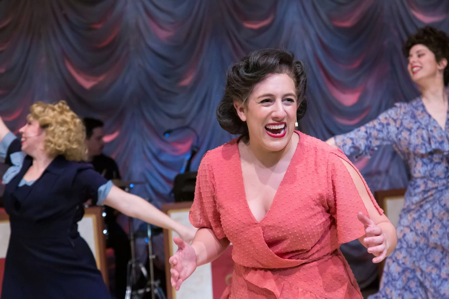 Several women dancing in a theater with a draped curtain backdrop, smiling and enjoying themselves.