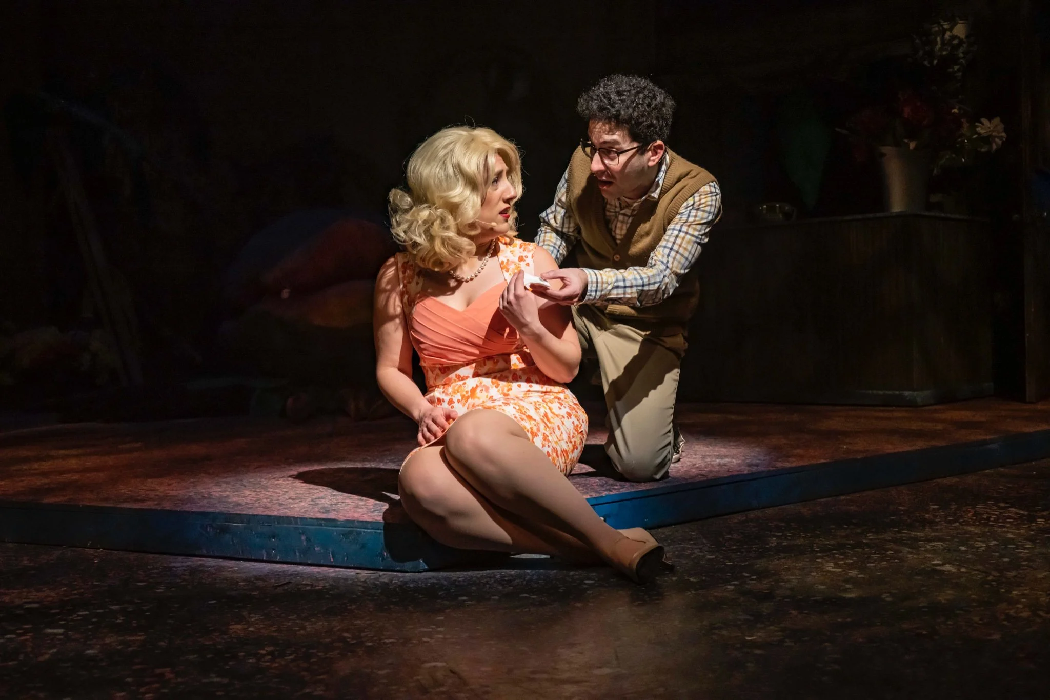 A theatrical scene with a woman in a floral dress sitting on the floor, speaking to a man in glasses and a checkered shirt who is kneeling next to her, holding her hand, in a dimly lit stage setting.