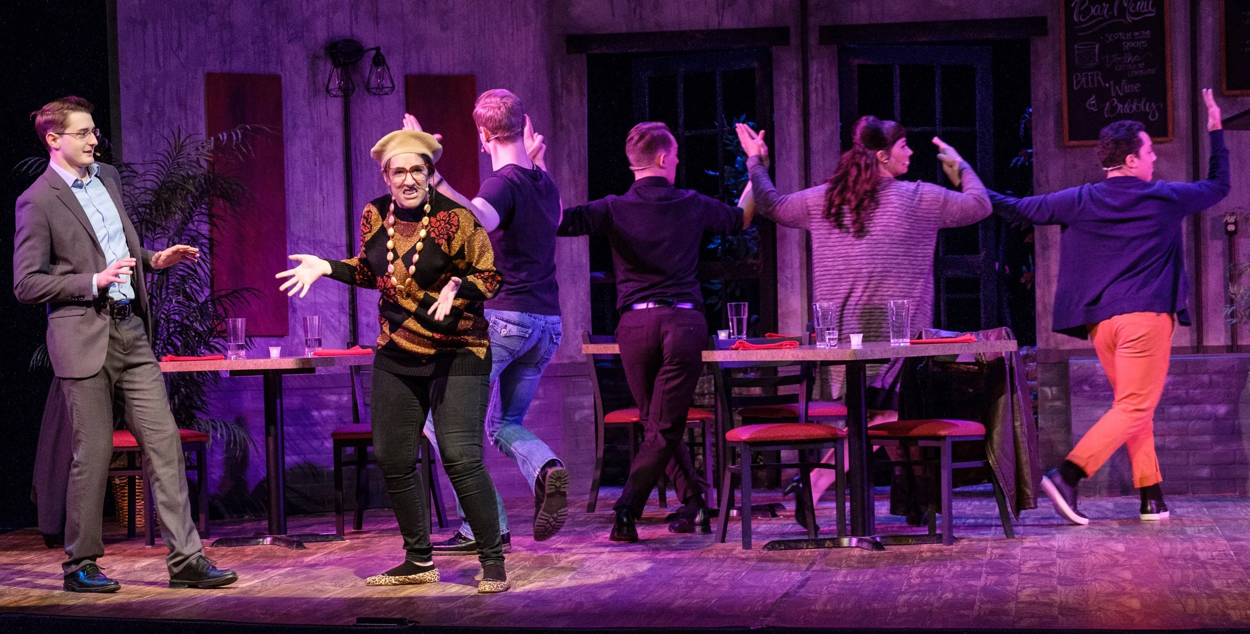Six people in a restaurant or cafe setting, engaged in a theatrical performance. The scene features a woman with glasses and a patterned sweater gesturing animatedly, while others are giving high-fives or interacting with each other. There are tables
