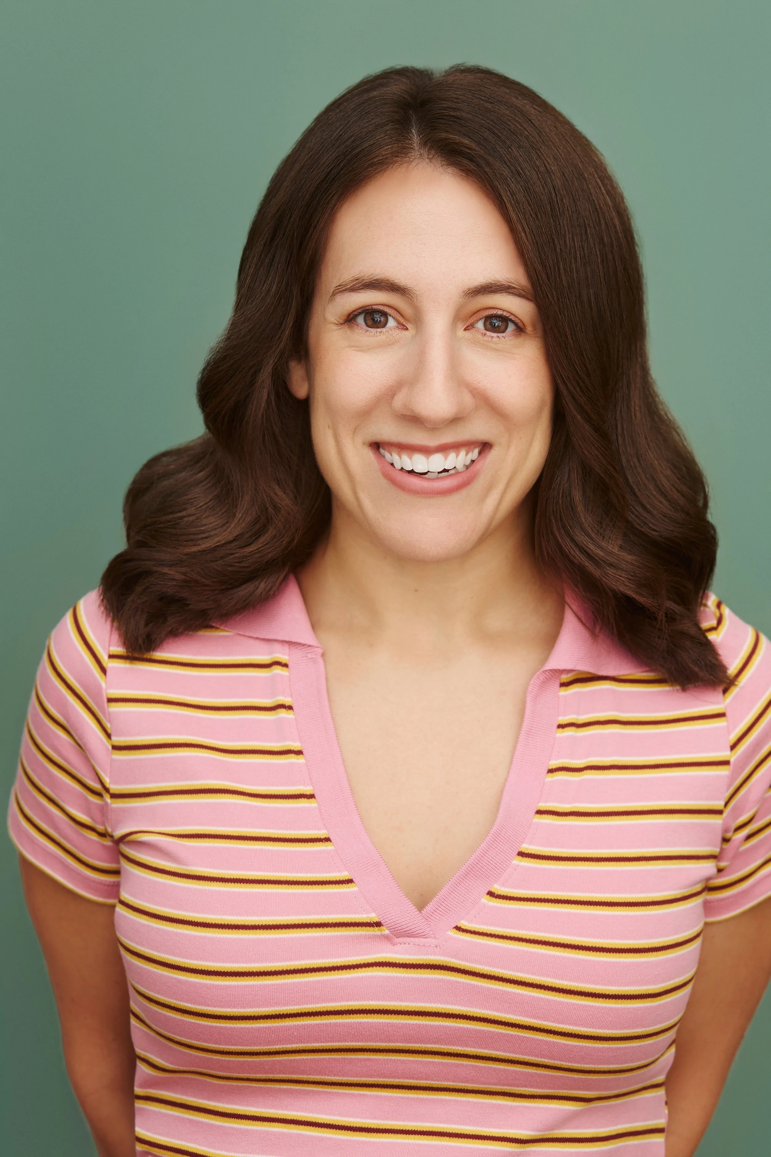 A young woman with shoulder-length brown hair wearing a pink and yellow striped shirt, smiling at the camera against a muted green background.