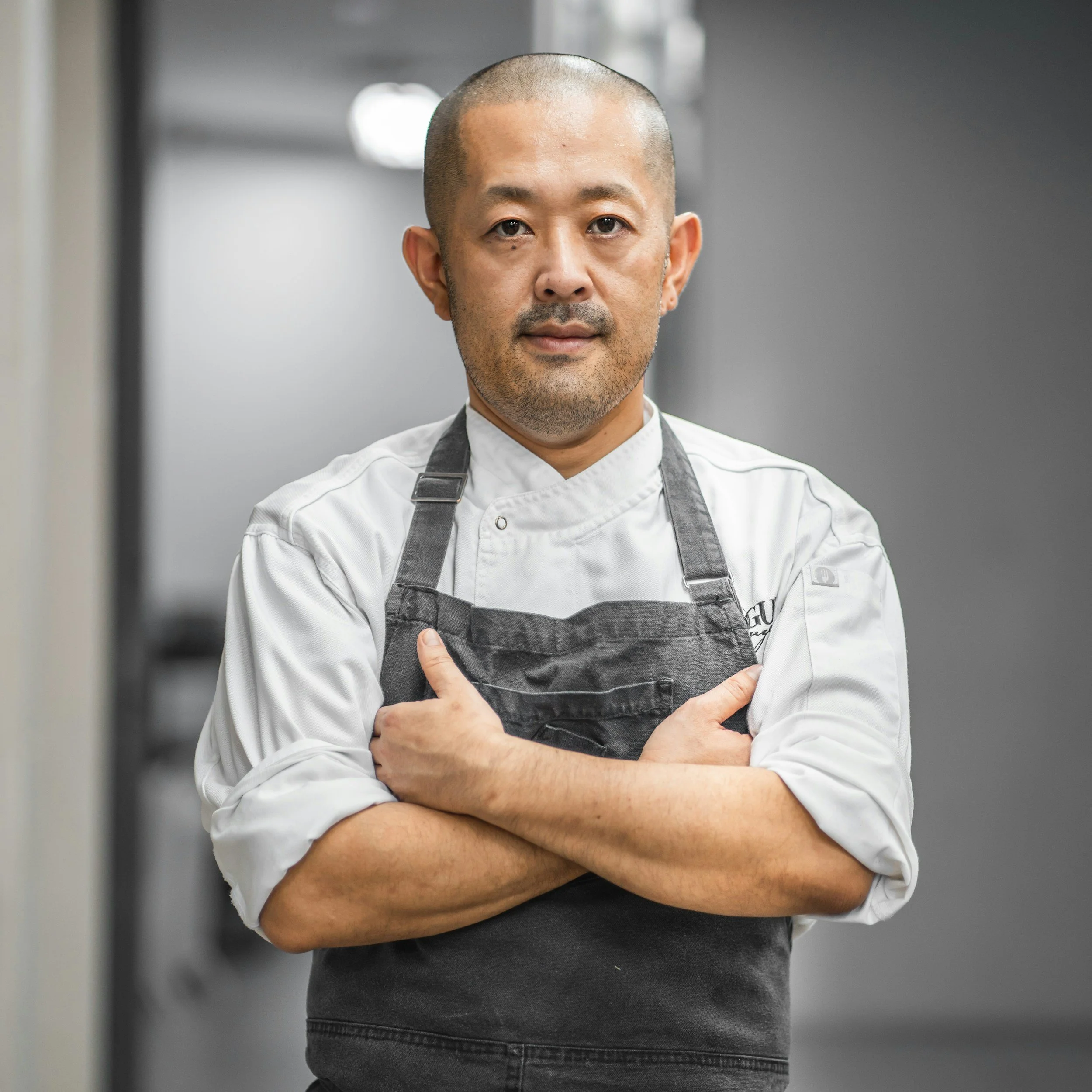 A male chef with short hair and a beard, wearing a white chef's coat and black apron, standing with arms crossed in a professional kitchen.