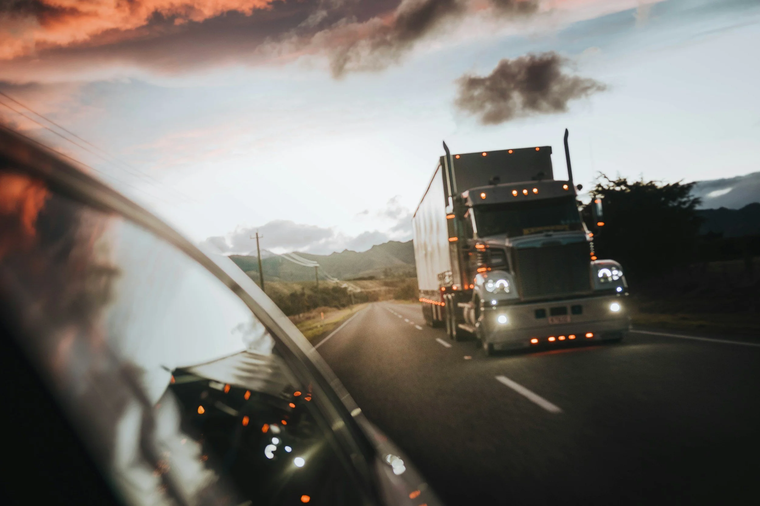 A semi-truck driving on a two-lane highway during sunset, with a mountain landscape in the background and a reflection of the truck's headlights in the side mirror.