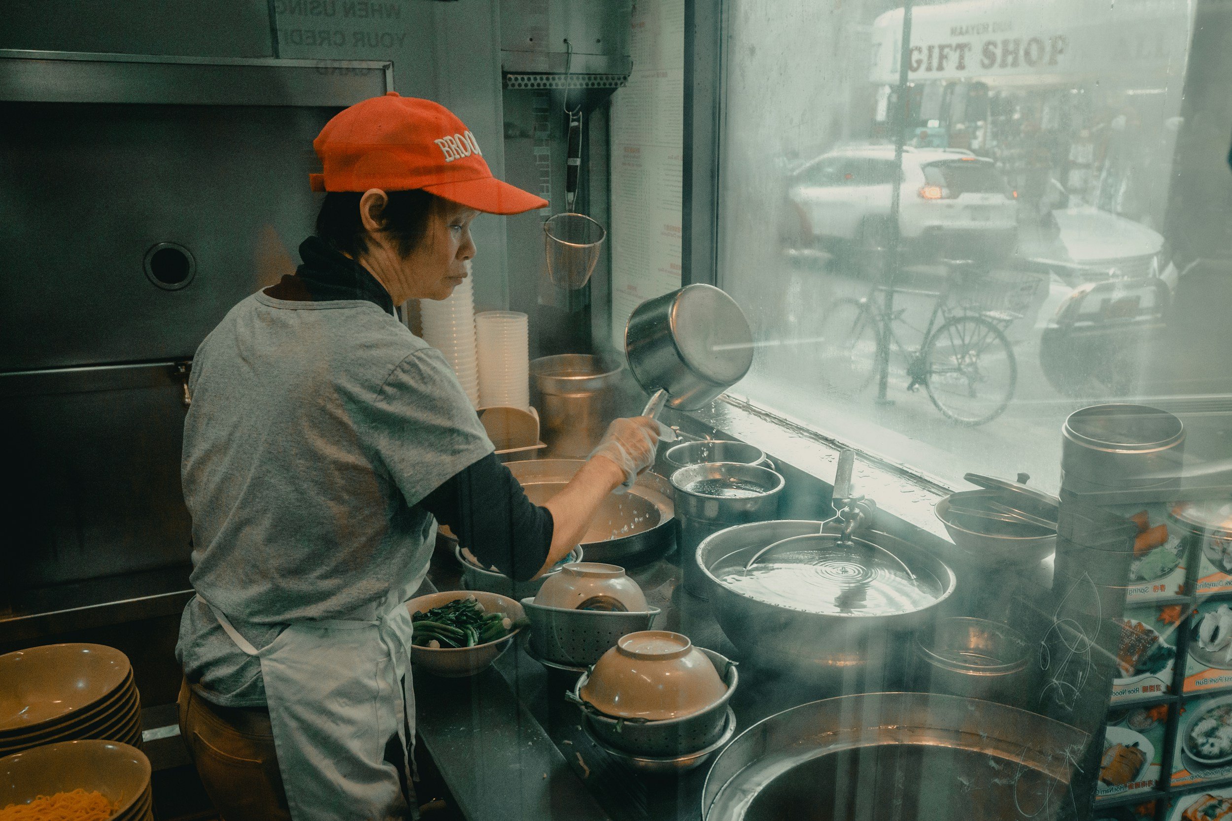 A woman in a red cap and apron prepares food in a small restaurant kitchen, with steam rising from a large pot and a window showing a street scene outside.