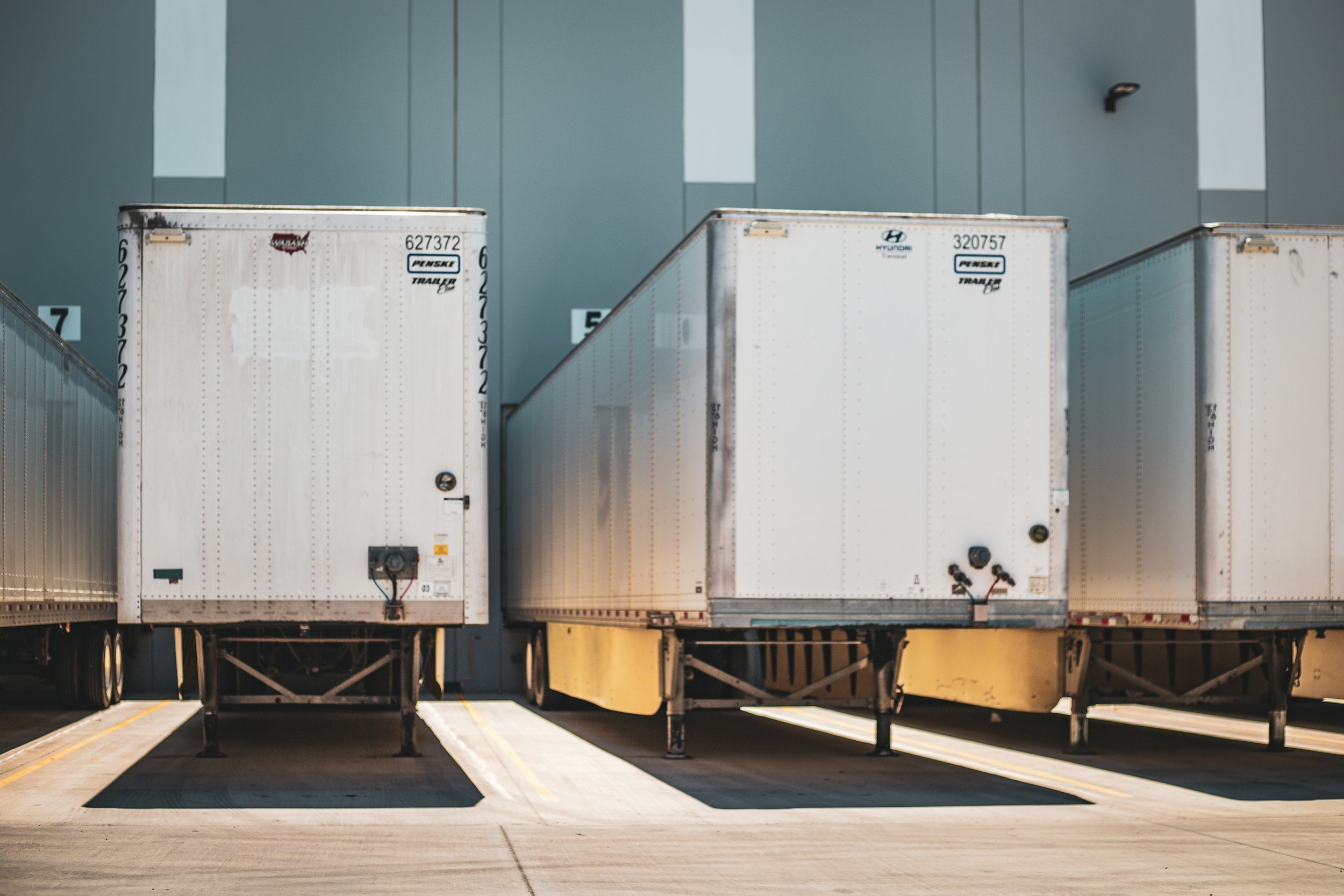 Three semi-trailers parked side by side in an outdoor lot with a large gray industrial building in the background.