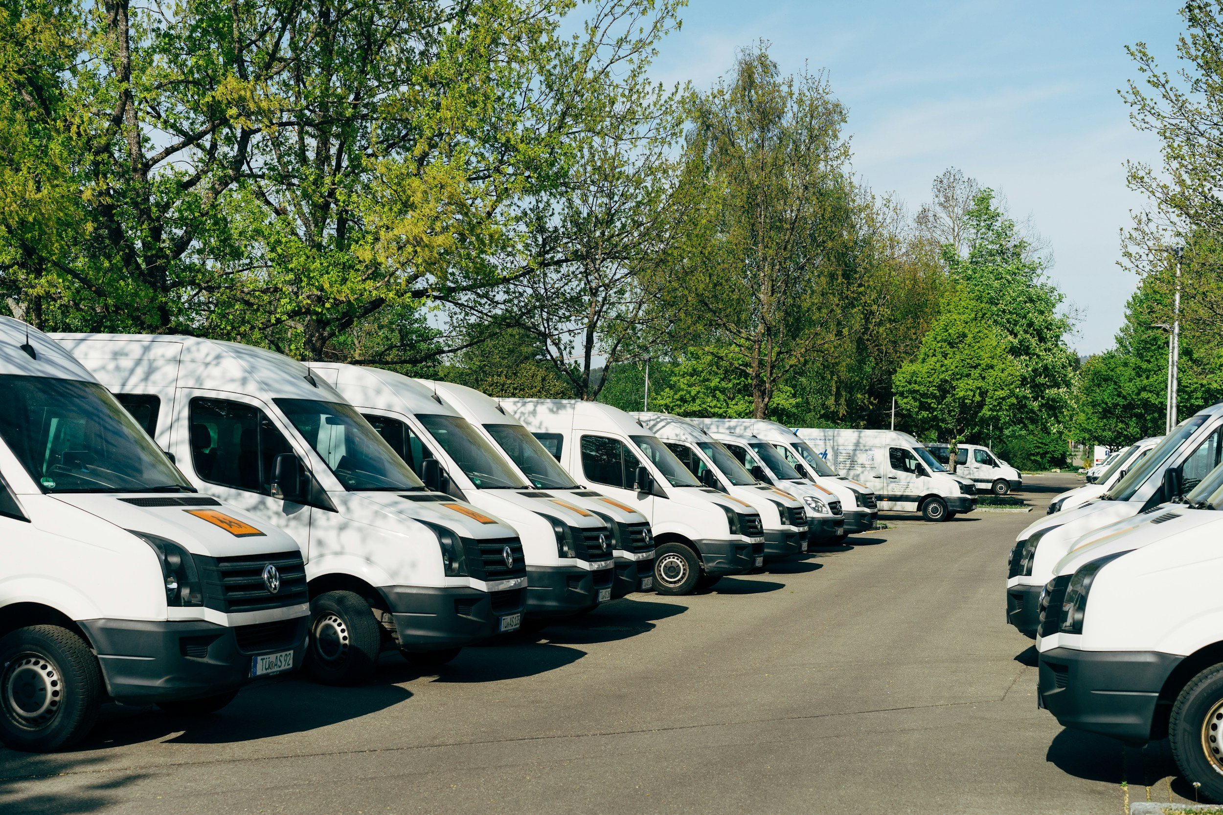 A parking lot filled with white commercial vans parked in a row under green trees on a sunny day.