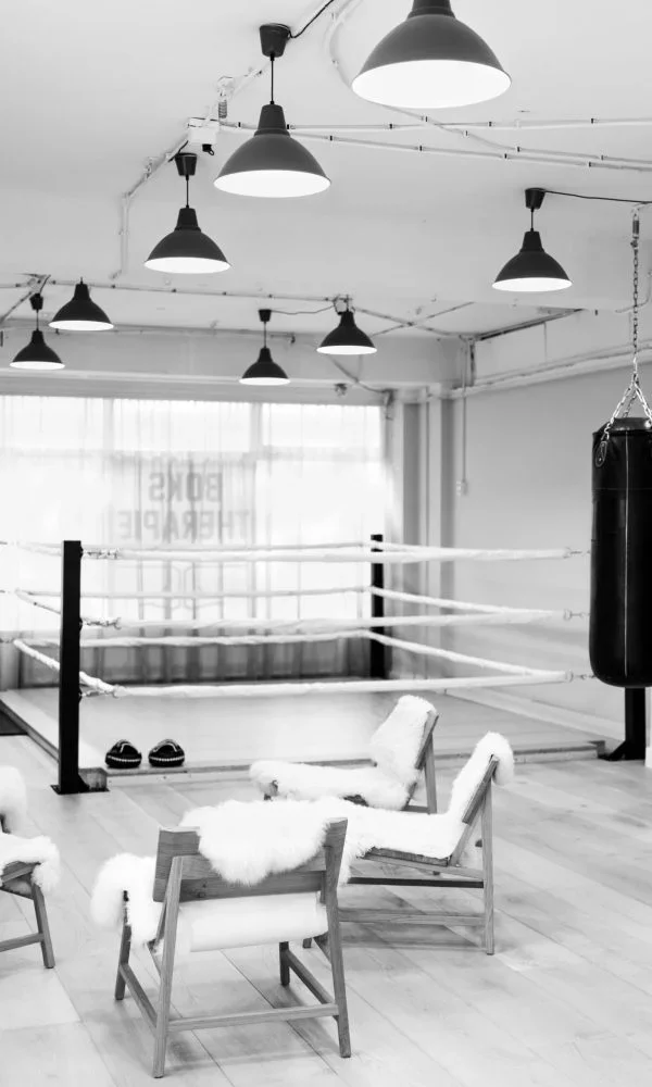 An empty boxing gym with hanging punching bag, boxing ring with ropes, and two chairs draped with fluffy white cloths in black and white.