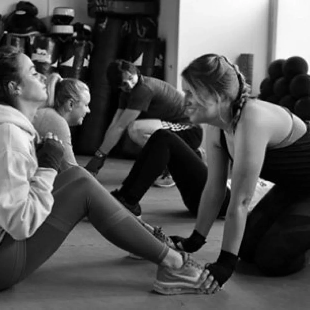 Women doing stretching exercises on the floor with a coach assisting, gym equipment in the background.