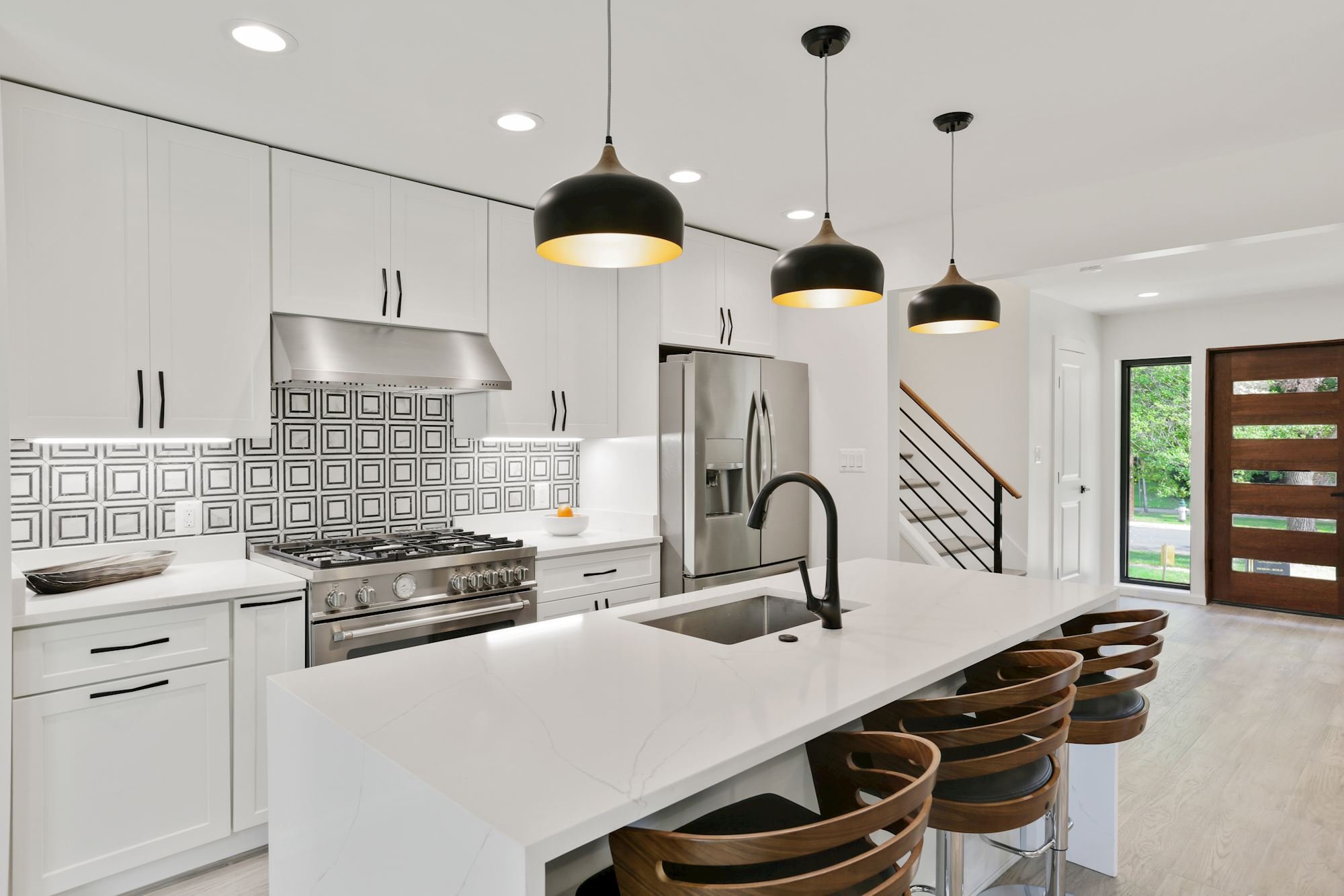 Modern renovated kitchen with white cabinetry, stainless steel appliances, black pendant lights, a white island with wooden bar stools, a decorative black and white tile backsplash, and large windows showing greenery outside.