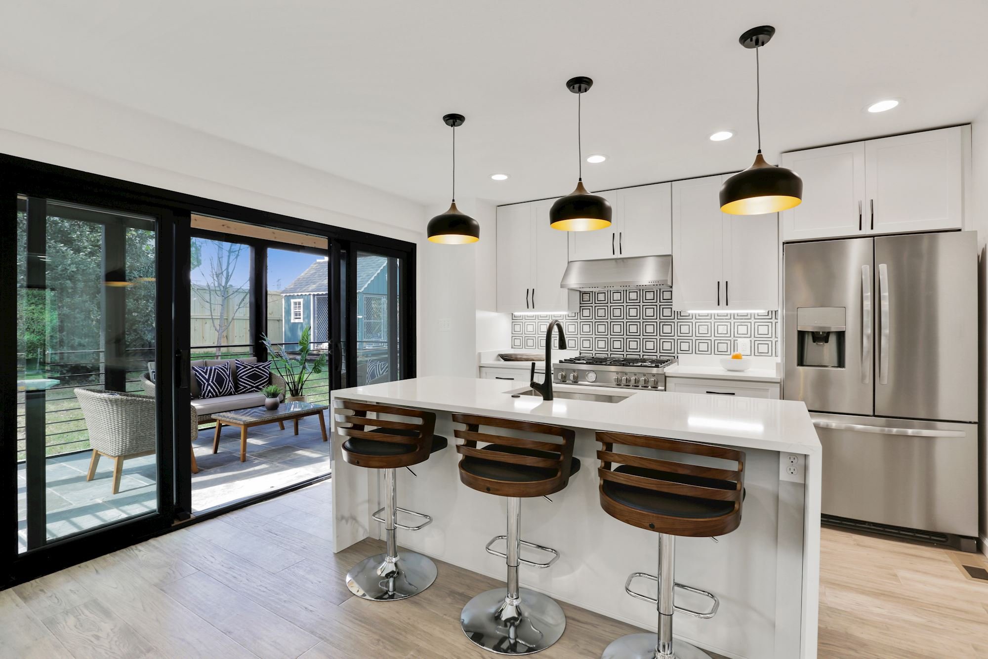 Modern kitchen renovation with white island and three wooden barstools, stainless steel refrigerator, black pendant lights, patterned backsplash, sliding glass door leading to outdoor patio with seating.
