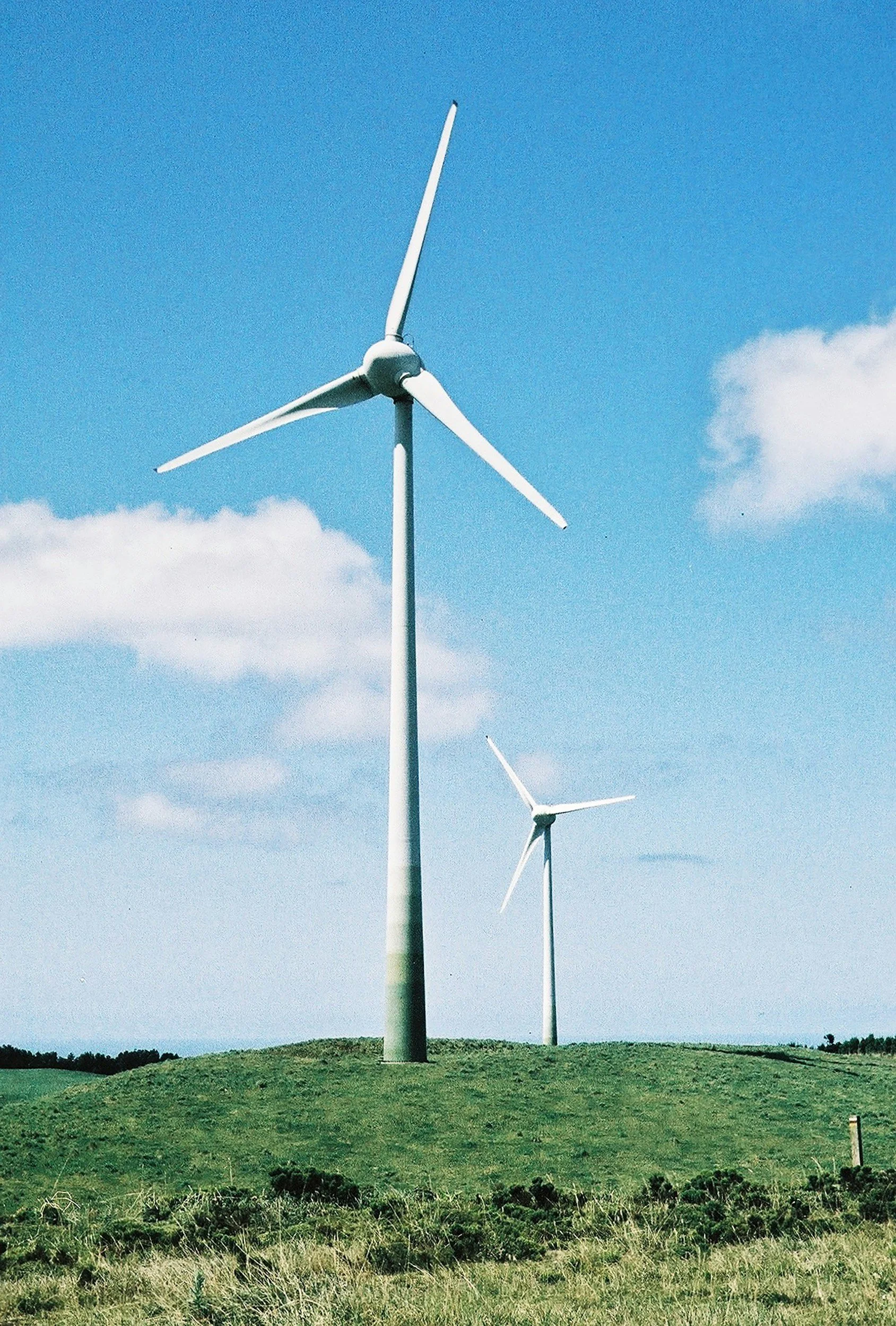 Two windmills sit atop a green hill with a blue, cloudy sky in the background.