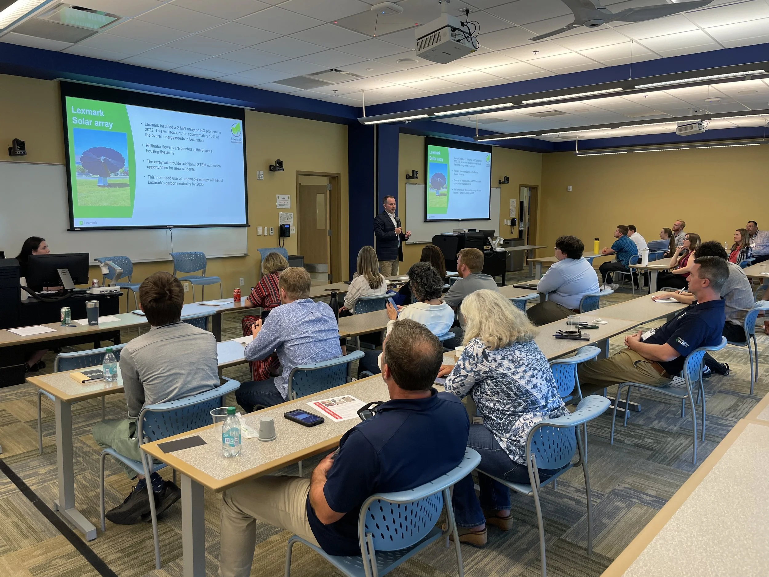 A group of people are seated at tables while listening to a man present a Home Energy Efficiency Workshop at the front of the room.