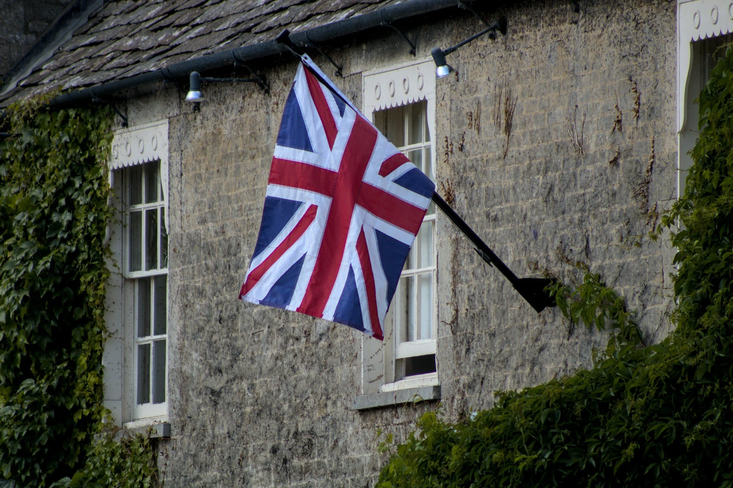 A British flag hanging on the exterior wall of an old stone building with ivy and greenery.