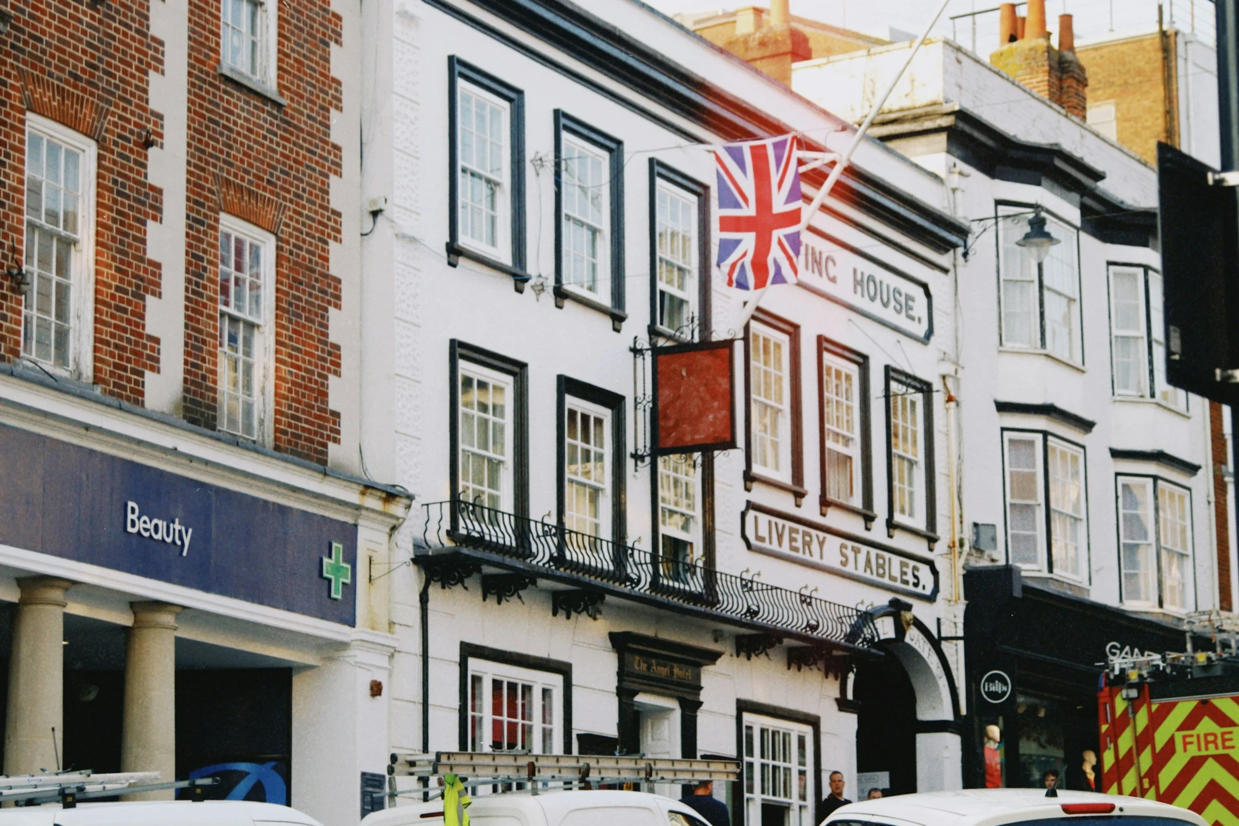 Street view with a building that has a Union Jack flag, signs for 'The Angel Hotel' and 'Livery Stables', and a fire truck parked outside.