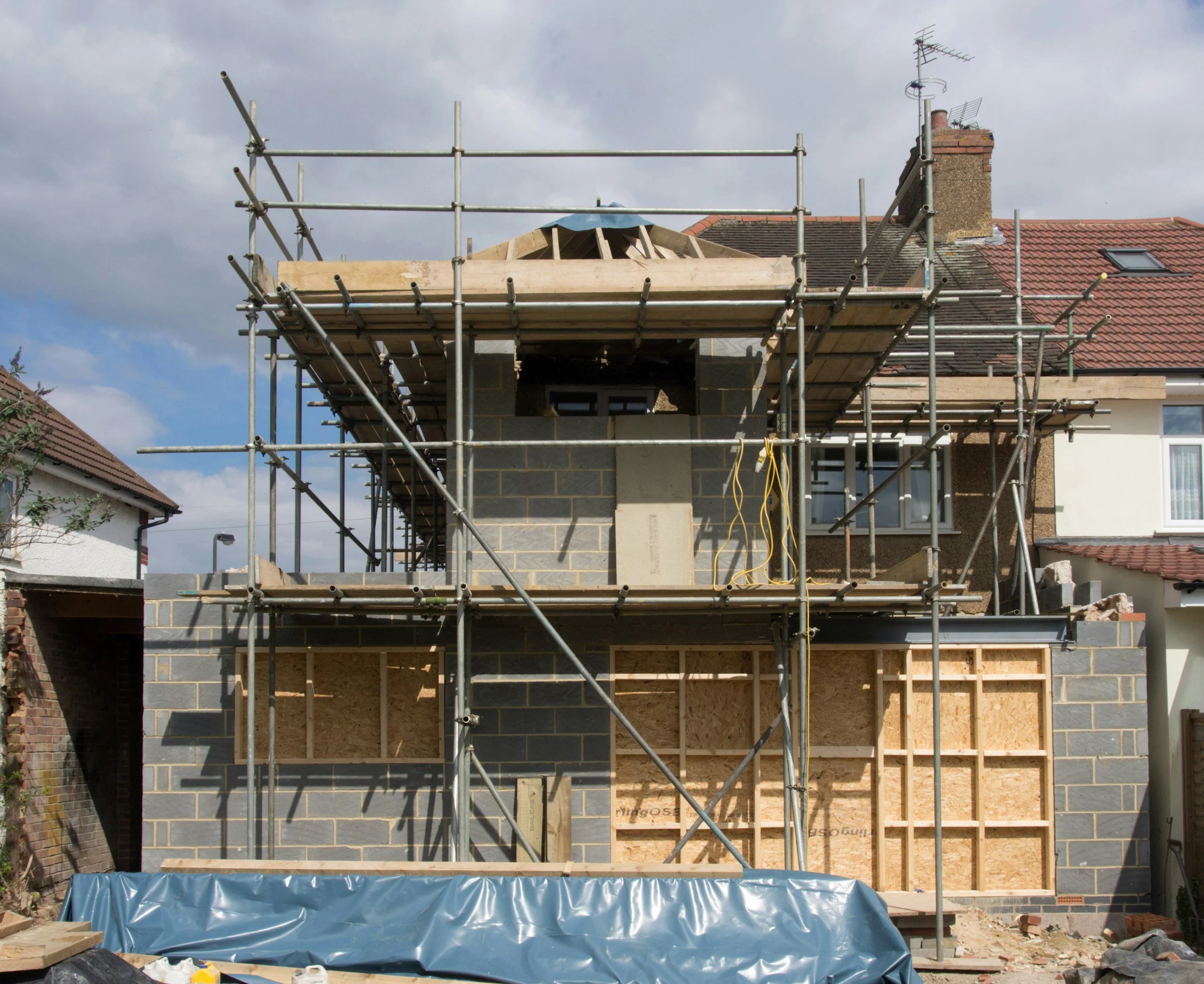 Under construction house with scaffolding around it, showing brick and wooden framing, on a sunny day with partly cloudy sky.