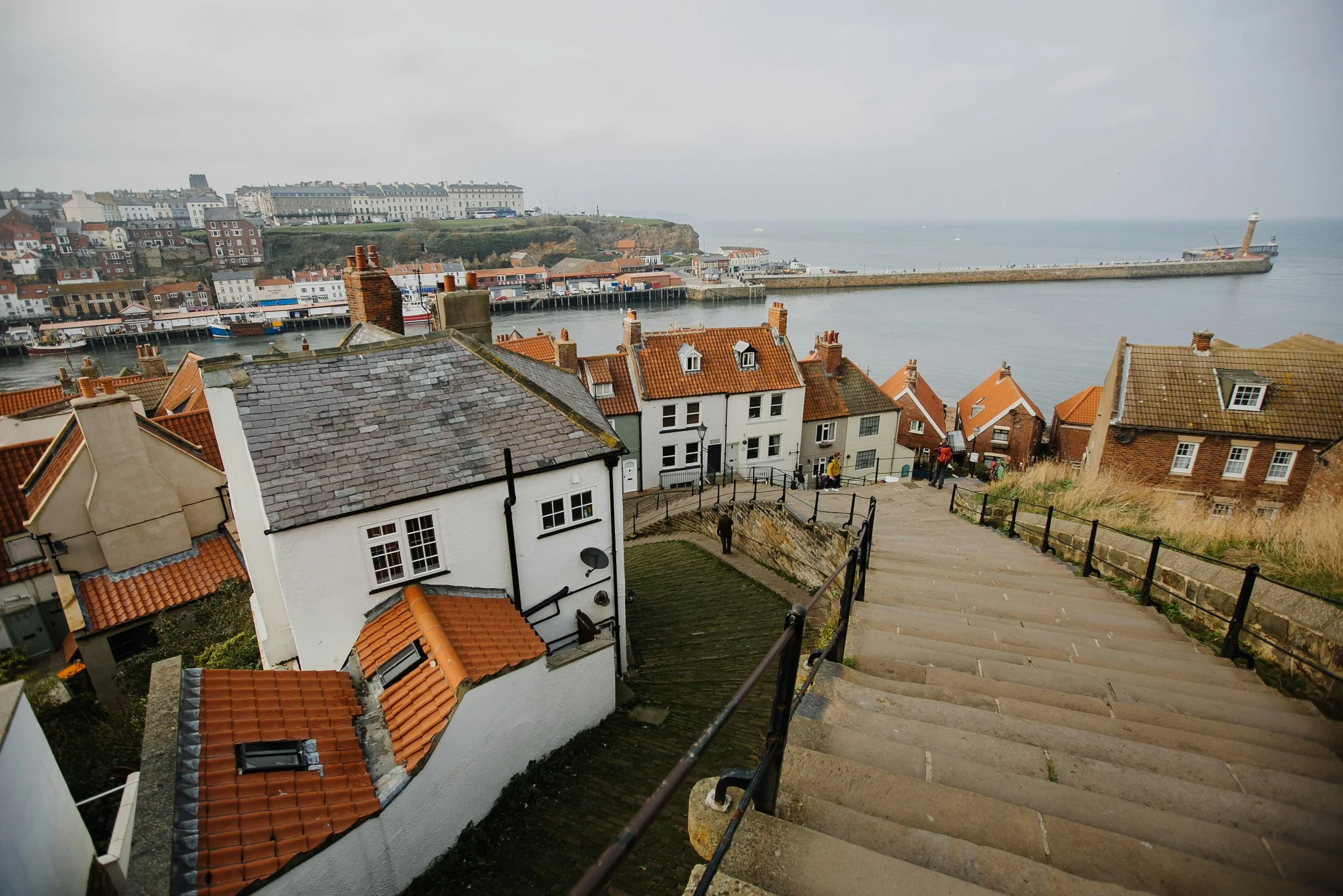 A coastal town with houses featuring red-tiled roofs, a steep stone staircase with black railings leading down, and a harbor with a breakwater and lighthouse in the background under a cloudy sky.