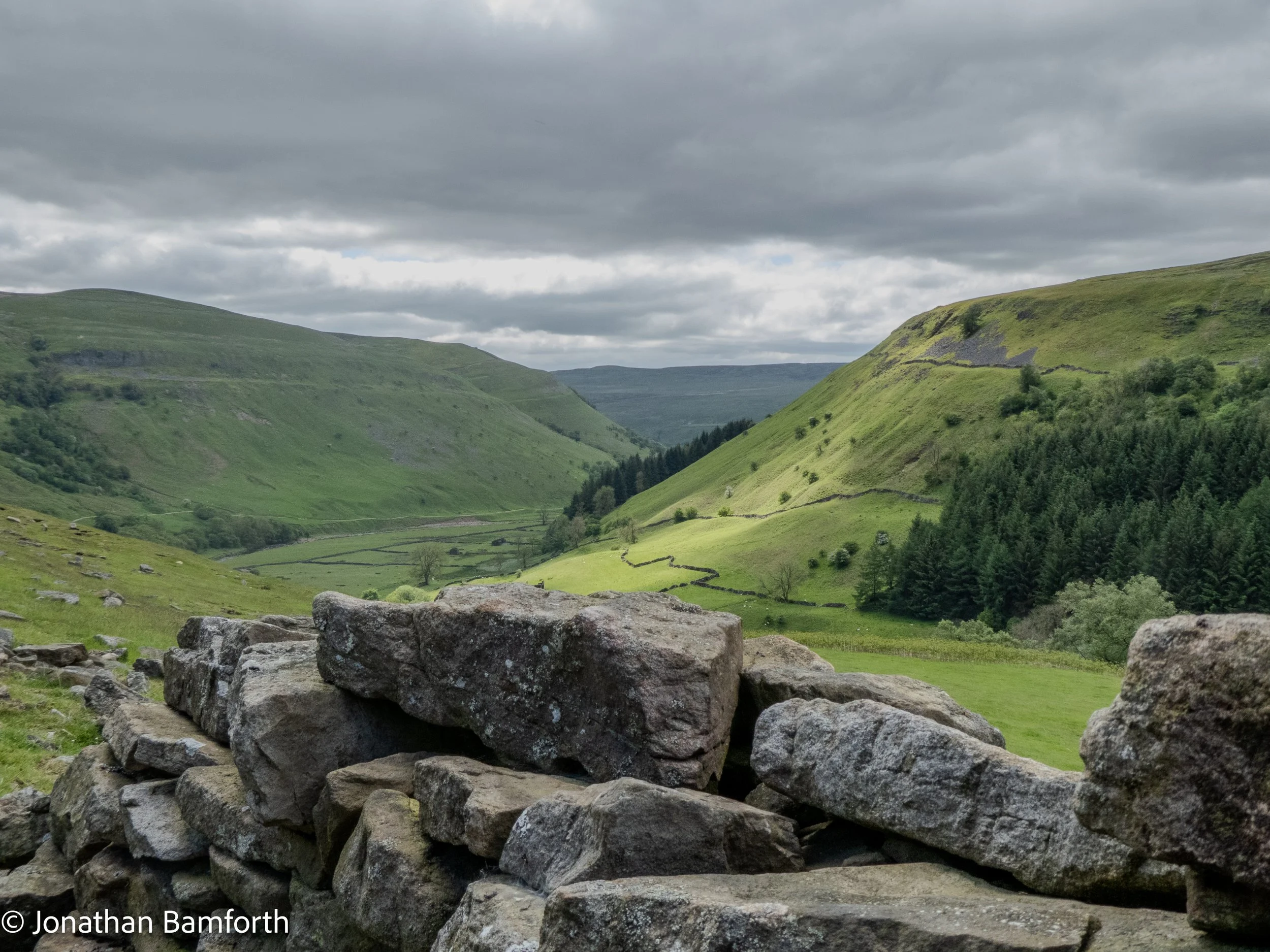 Scenic view of lush green hills and valley, with a stone wall in the foreground and cloudy sky overhead.