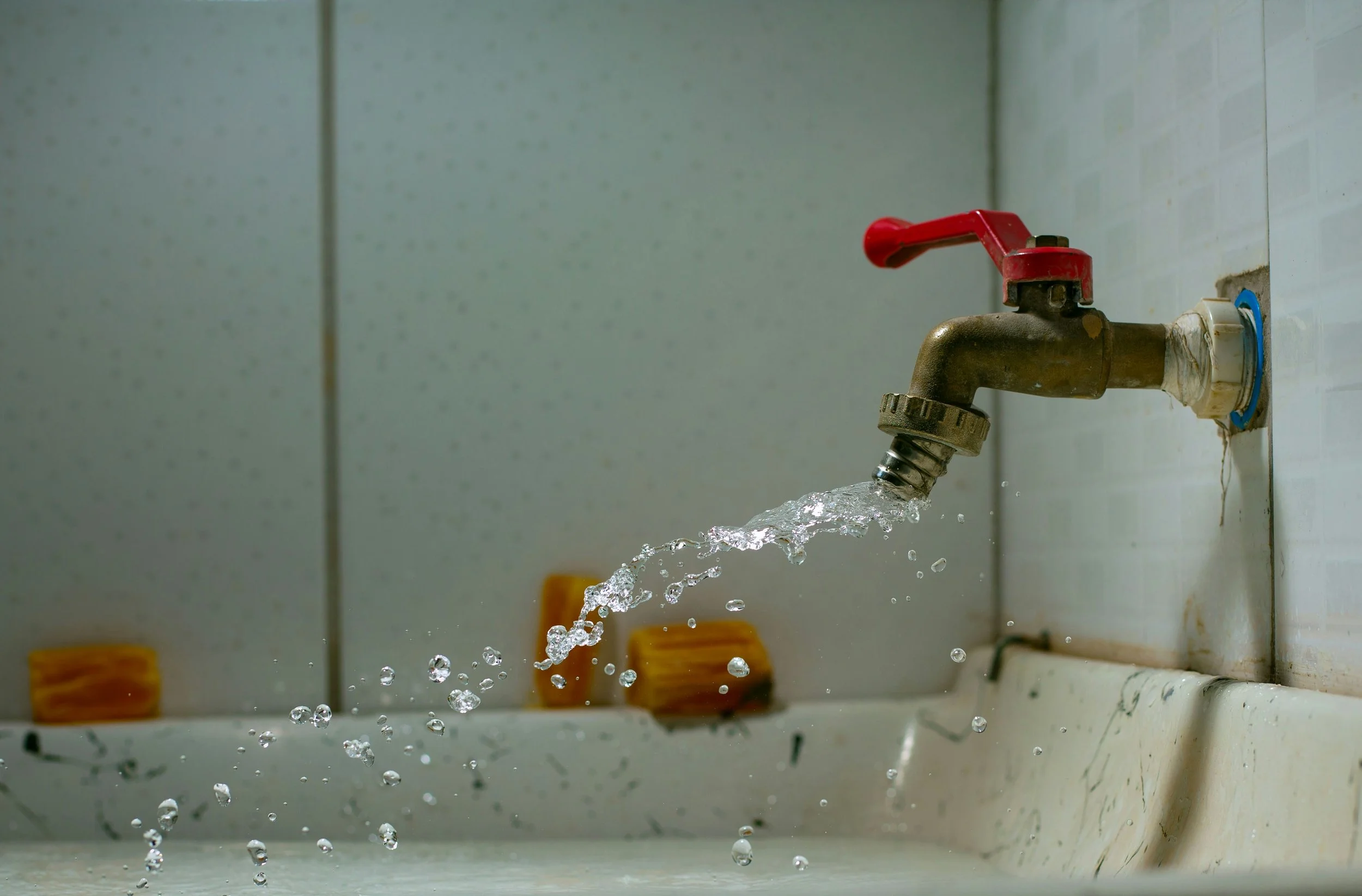 A close-up view of a rusty faucet with a red handle, turned on, with water flowing into a white sink, in a tiled room.