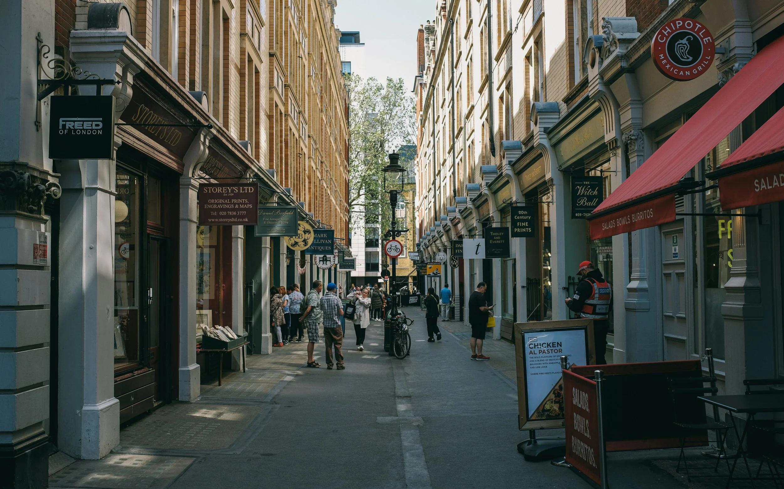 A busy urban street with shops on both sides, pedestrians walking, and outdoor signs advertising various stores and restaurants.