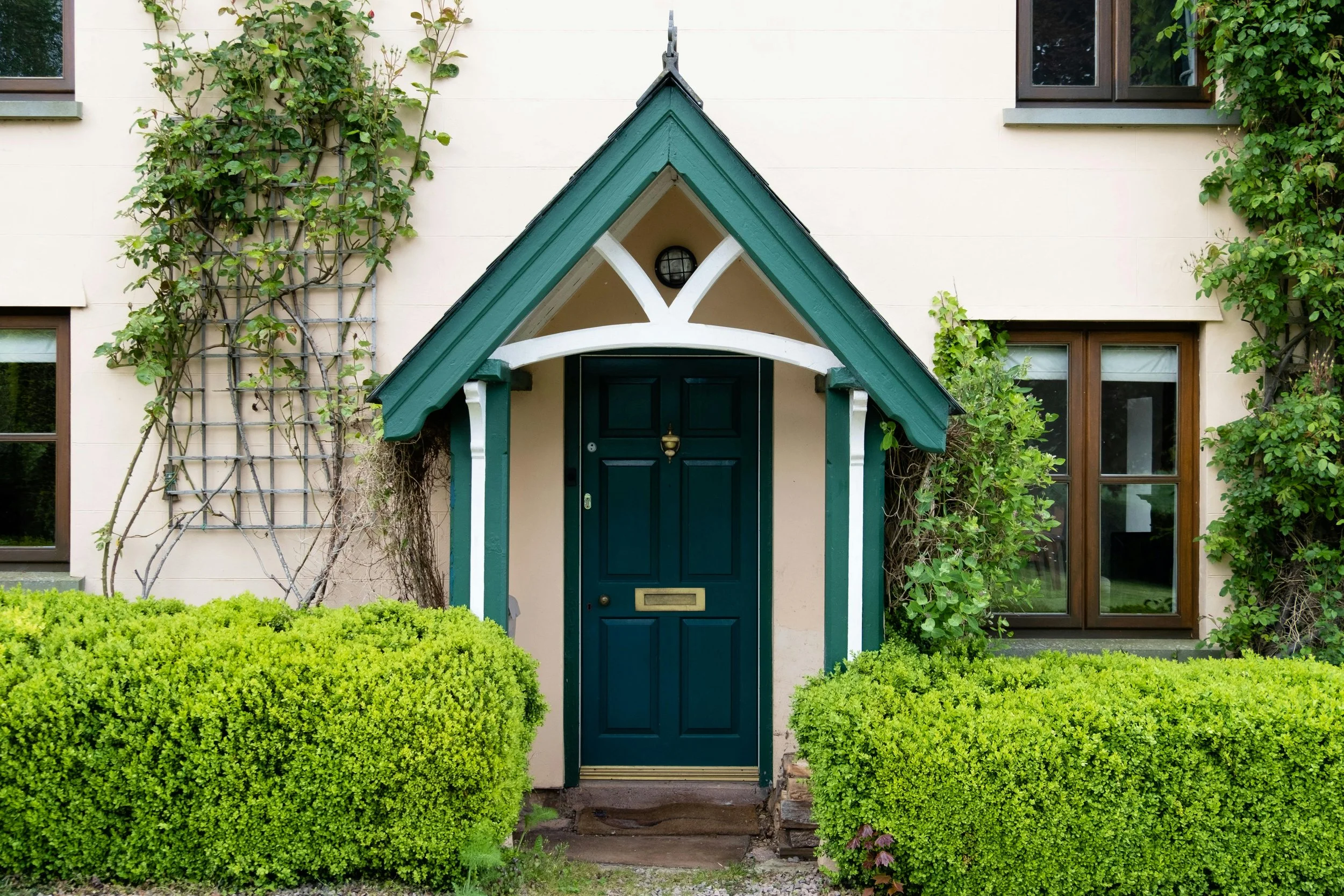 Front entrance of a house with a dark green door, small gabled roof, surrounded by lush green bushes and climbing plants, with windows on either side.