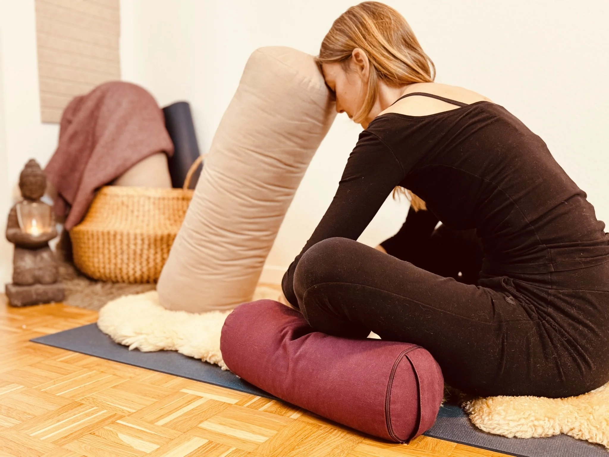 Frau beim Yoga, in einem Raum mit Holzfußboden, sitzend auf einer Yogamatte, mit Kissen und einem Nackenkissen.
