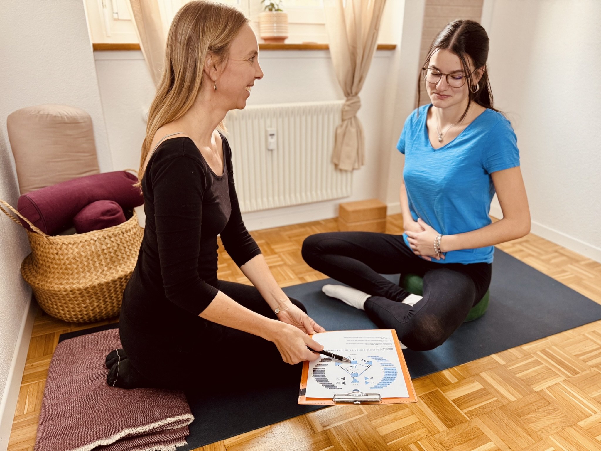 Zwei Frauen bei einer Yoga- oder Meditationssitzung im Wohnzimmer, eine zeigt auf einen Plan auf einem Clipboard.