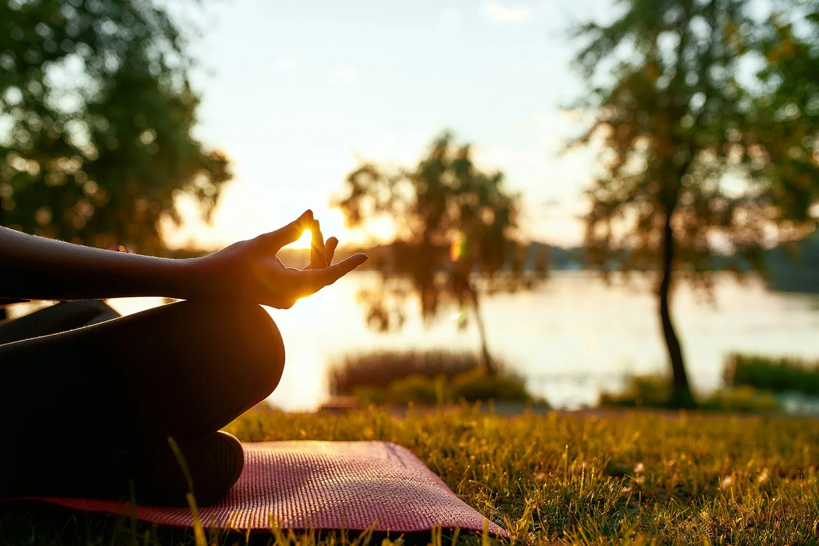 Nahaufnahme einer Person, die im Lotussitz meditiert, mit Sonnenuntergang im Hintergrund an einem Wasserufer