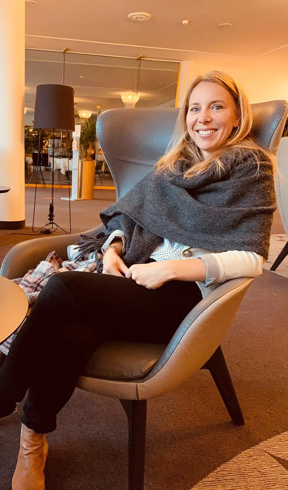 Eine lachende Frau sitzt in einem modernen Sitz im Hotel oder Restaurant, im Hintergrund sind Pflanzen, Lampen und ein Fenster sichtbar.