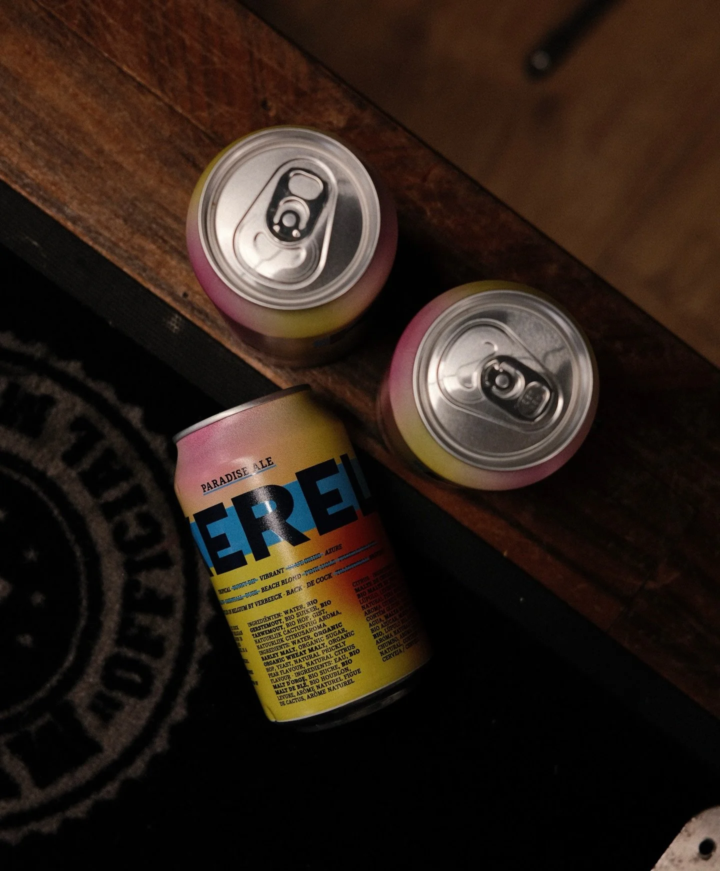 Deux canettes de boisson à côté d'une canette plus grande, posé sur un tapis noir avec un motif, sur un meuble en bois.