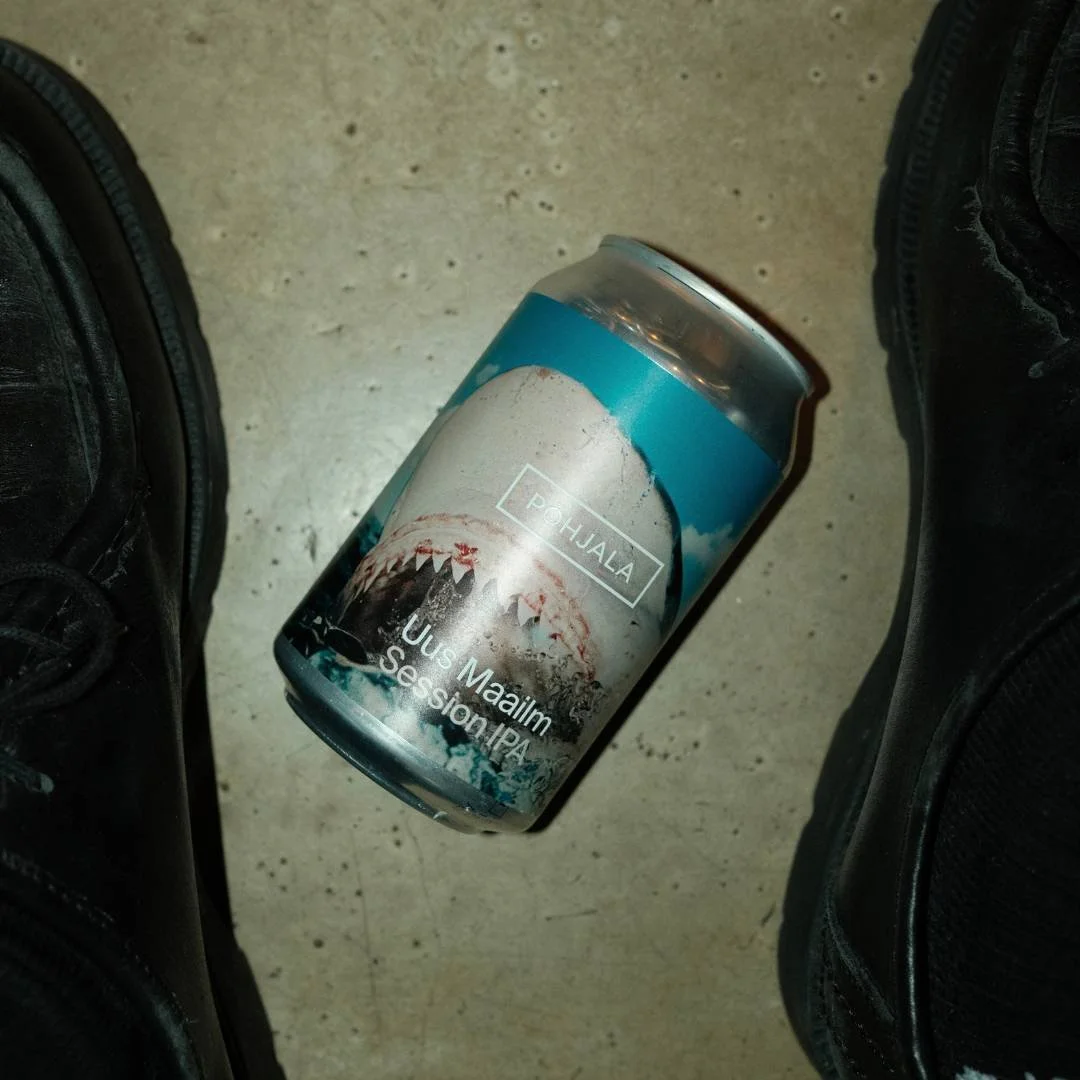 Bière en canette avec un paysage de montagnes et de nuages sur l'étiquette, posée au sol entourée de chaussures noires.