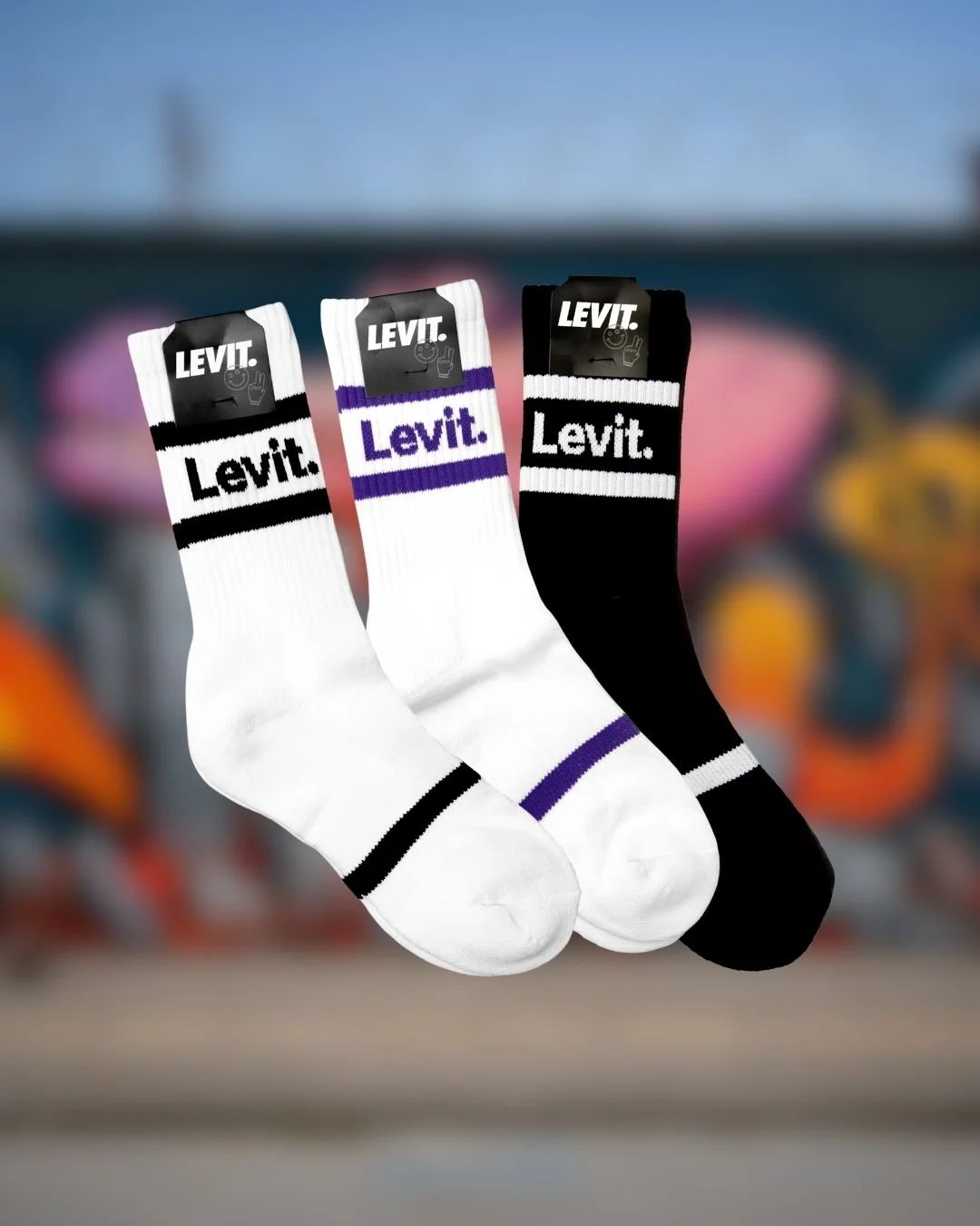 Trois paires de chaussettes de sport Levi's en blanc, noir et blanc avec rayures, et blanc avec rayures violettes, suspendues. Fond flou avec des objets colorés et sportifs.