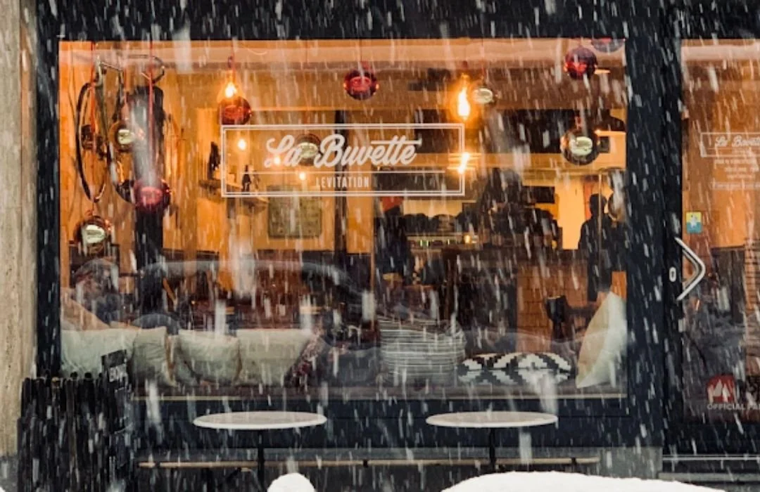 Vitrine d'un café ou restaurant appelé La Buvette, vue de dehors sous une forte pluie, intérieur chaleureux avec clients, décor industriel avec luminaires suspendus.