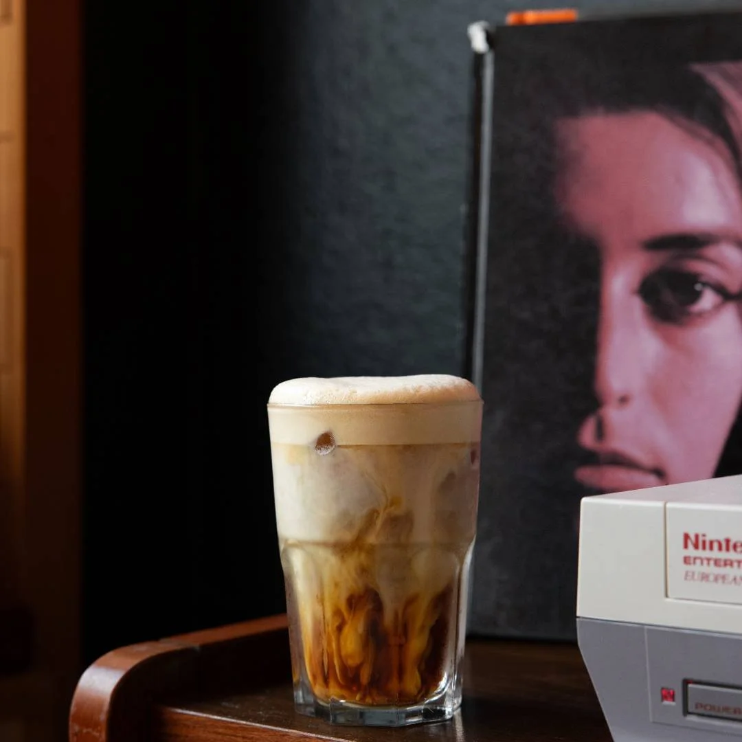 Verre de café glacé avec de la crème et du caramel sur une table en bois, avec un poster d'une femme sur le mur en arrière-plan.