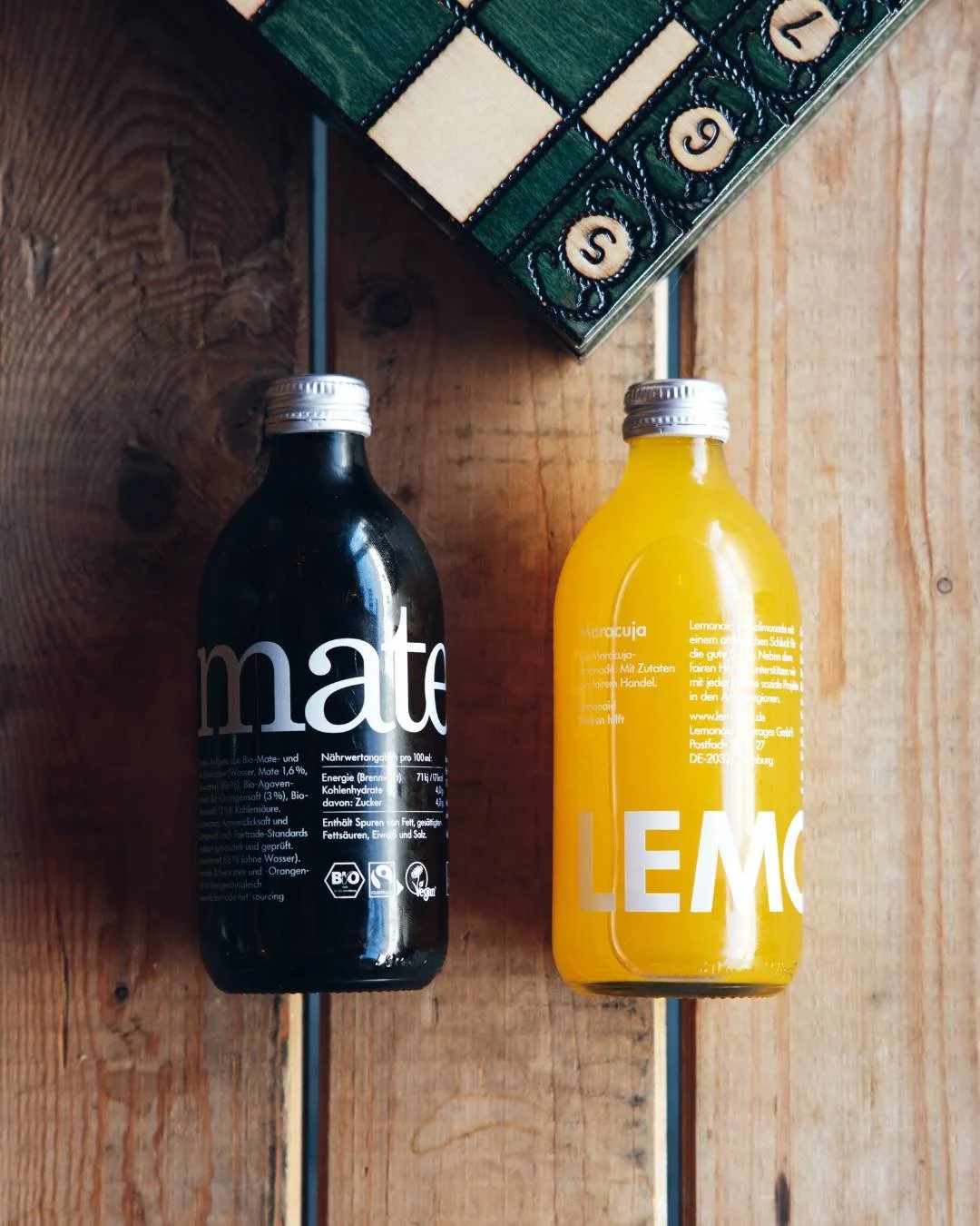 Deux bouteilles, une de boisson noire appelée Mate et une de boisson jaune appelée Lemonade, posées sur une surface en bois avec un jeu de société en arrière-plan.