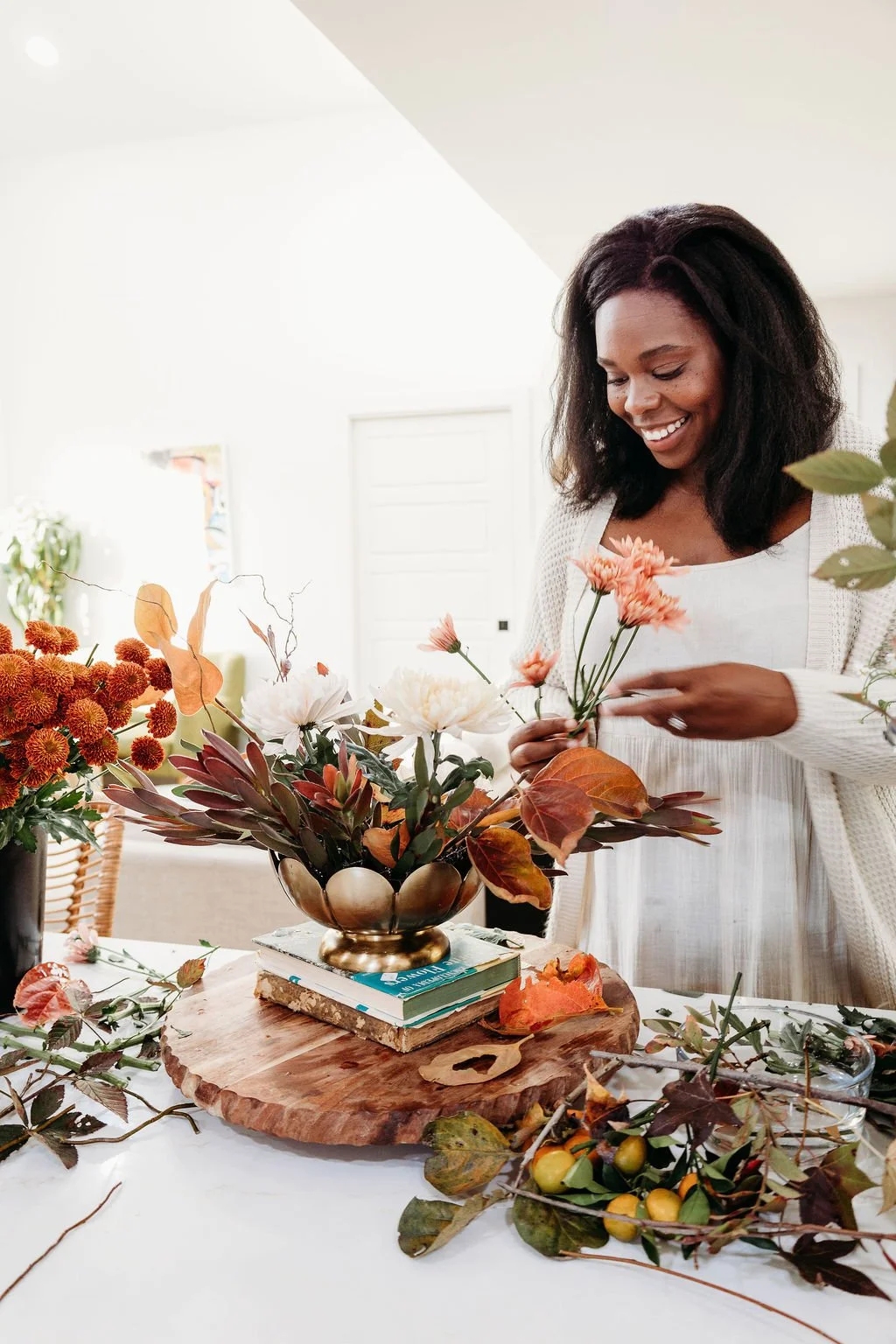 A woman arranging flowers at a table with various autumn leaves, branches, and floral arrangements around her.